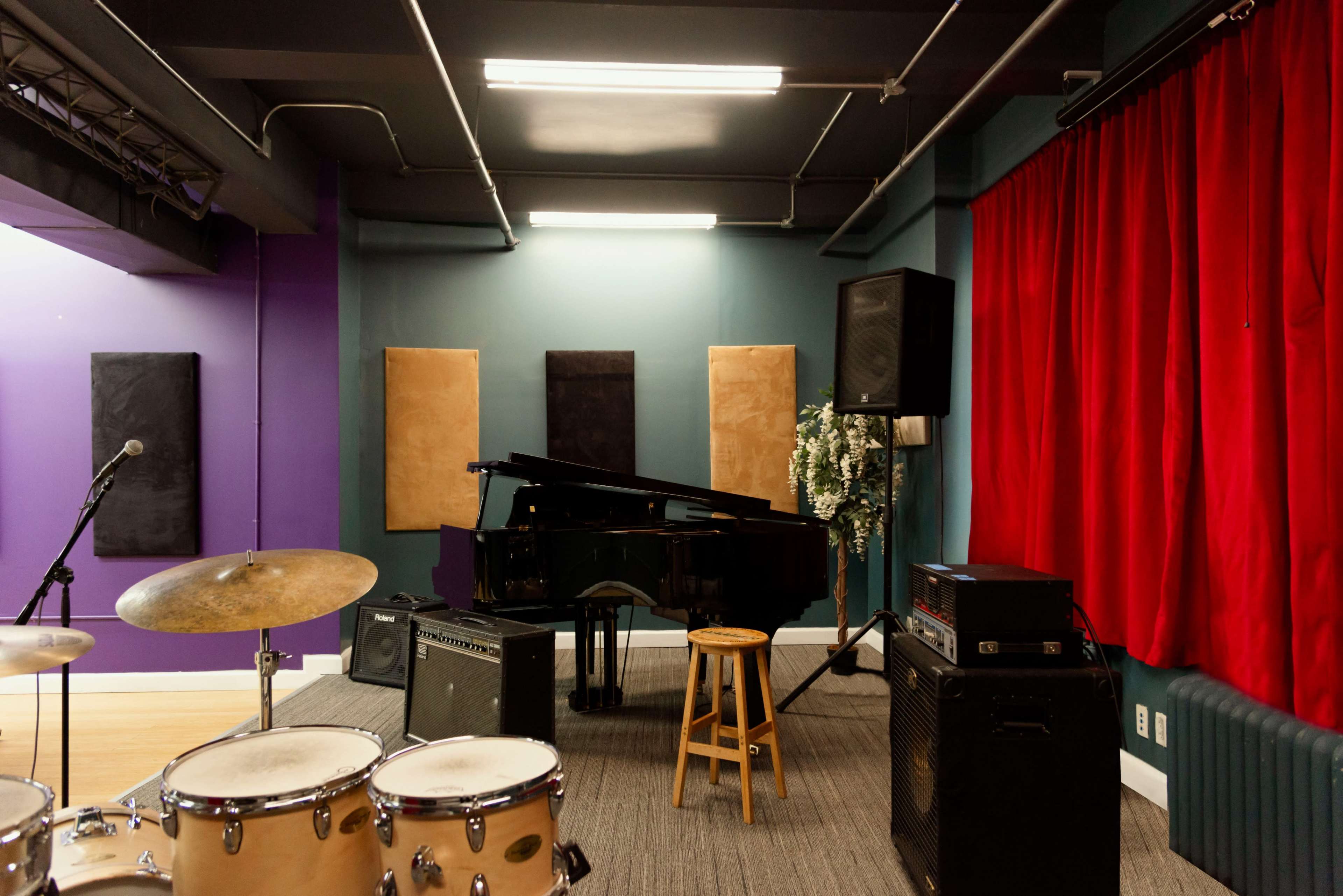 The image shows a music rehearsal studio with a grand piano in the center, surrounded by various musical instruments and equipment, against a backdrop of colorful walls and red curtains.