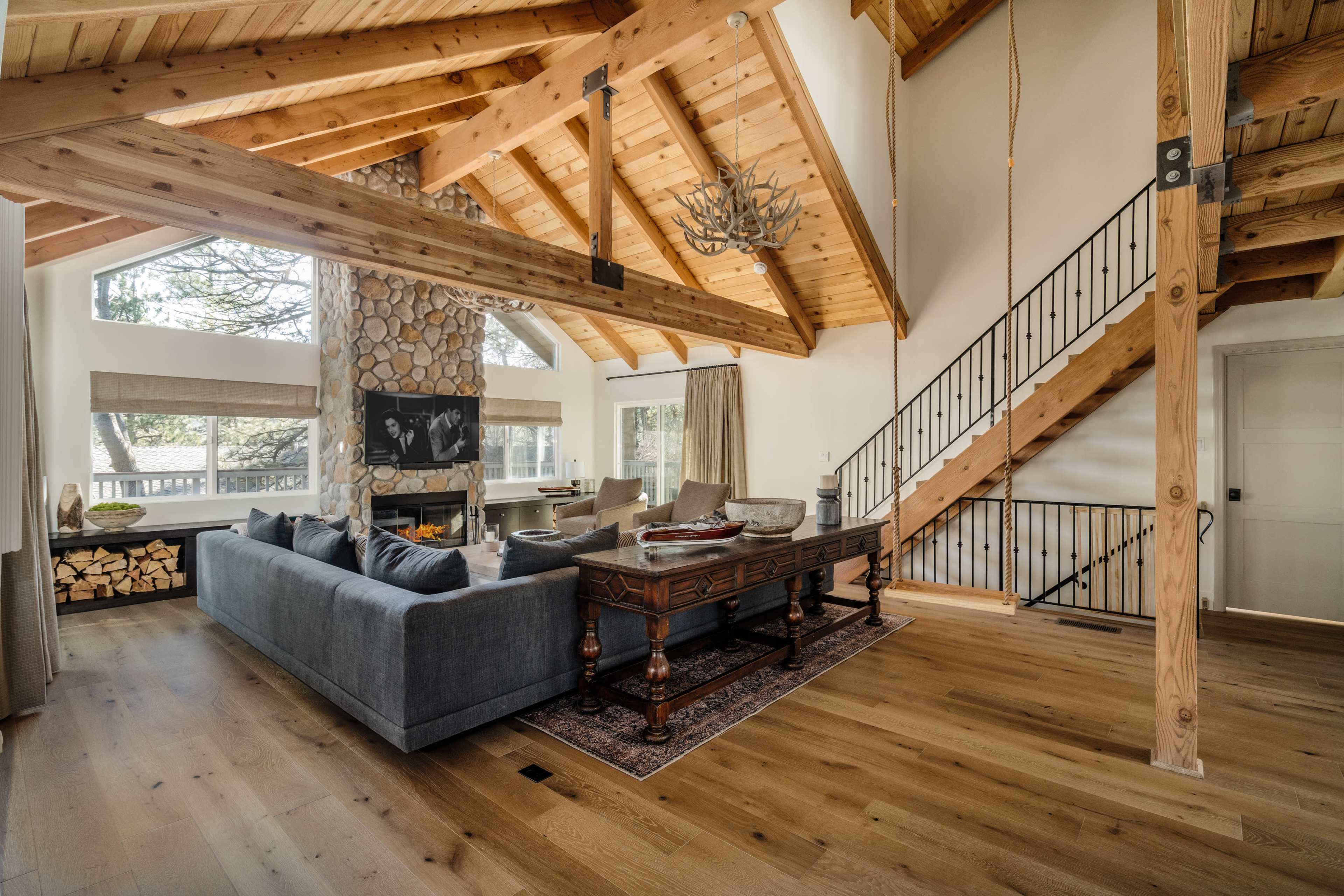 The interior of a spacious living room features wooden beams, a stone fireplace, a large gray sectional sofa, and a staircase leading to an upper level.