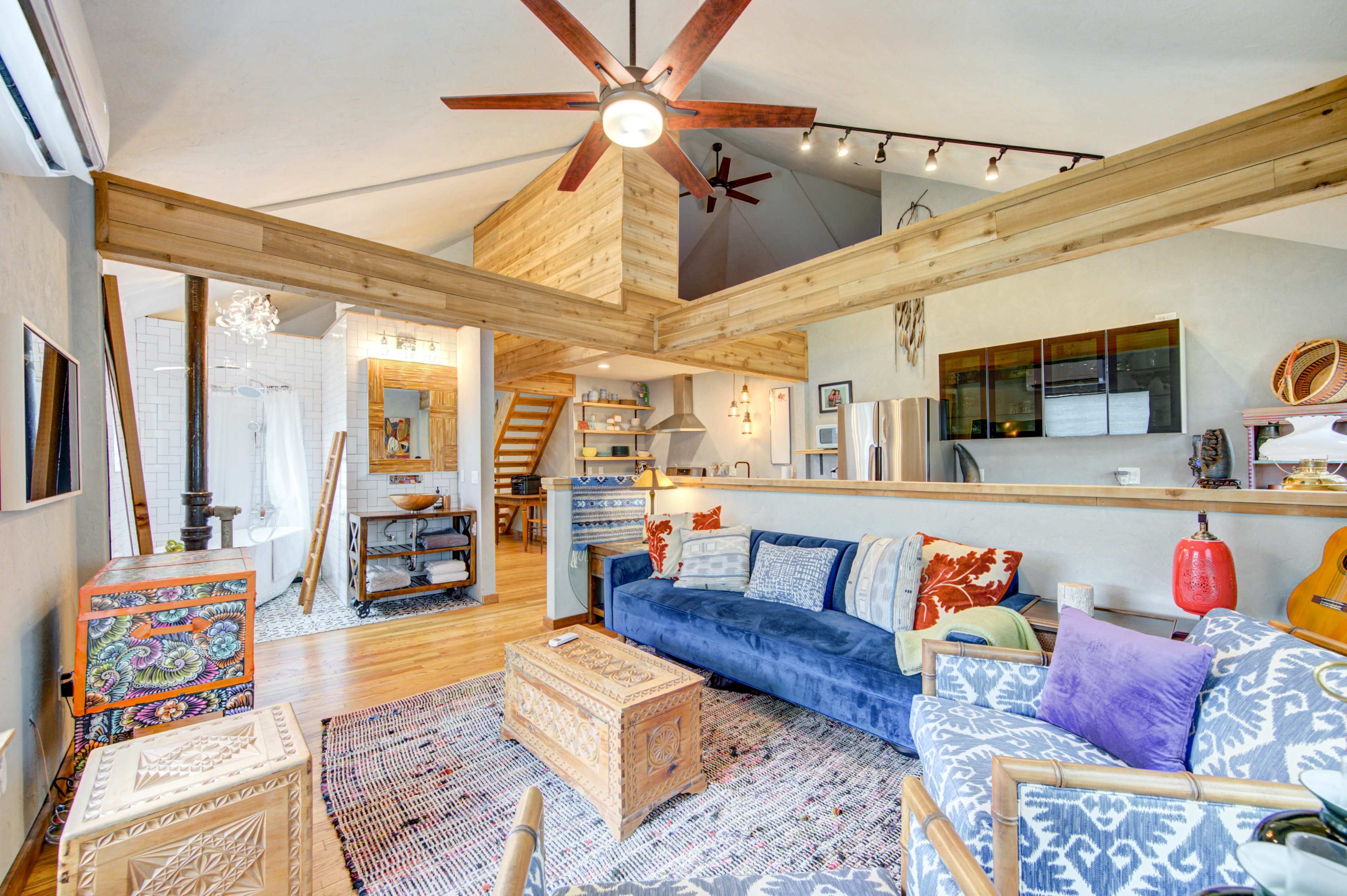 A modern, open-concept living area featuring a blue sofa, a wooden ceiling with ceiling fans, and decor that includes a patterned rug and various furnishings.