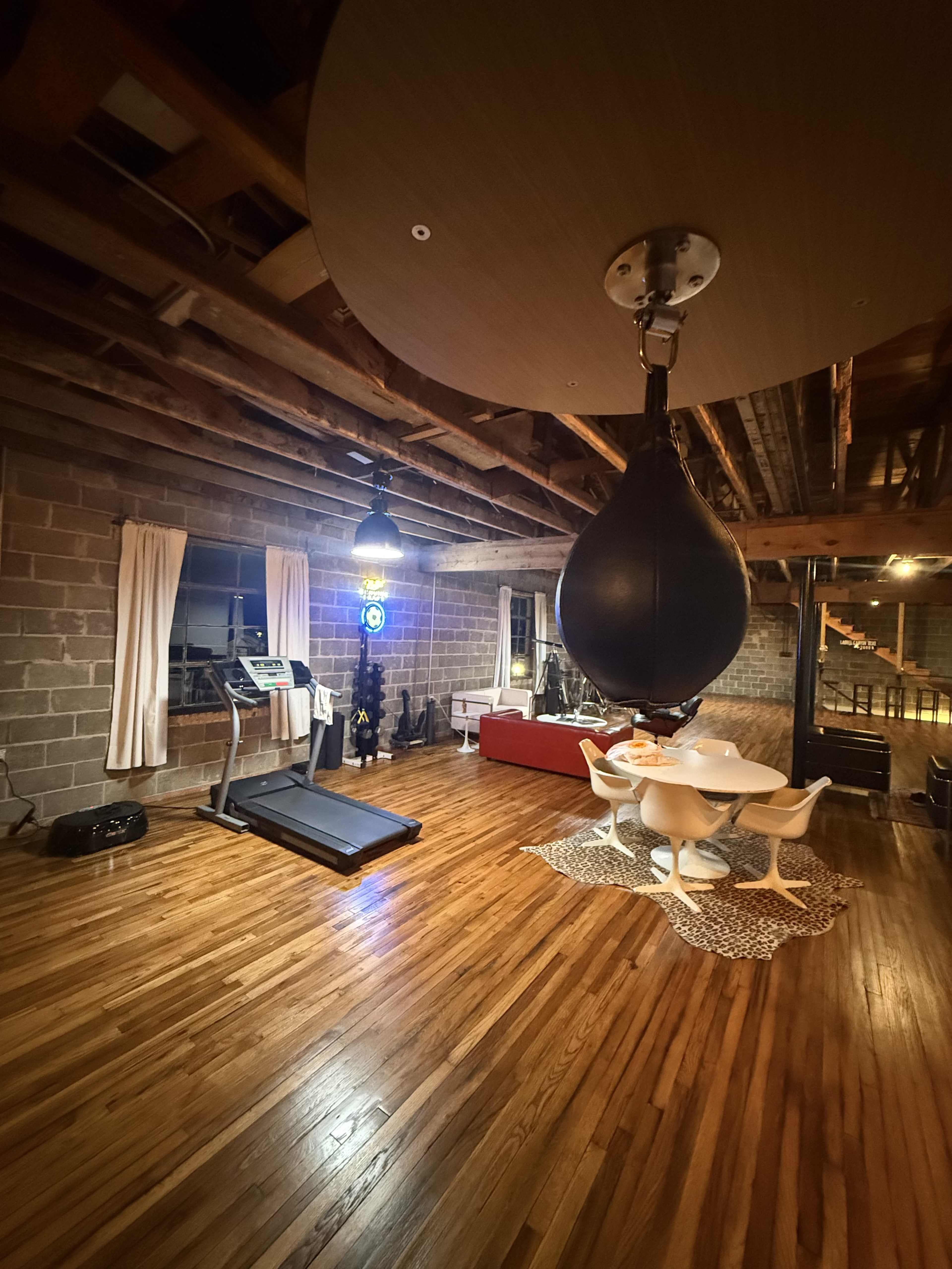 The image shows a spacious room with wooden floors featuring a treadmill, a punching bag hanging from the ceiling, a red sofa, and a round dining table set under soft lighting.