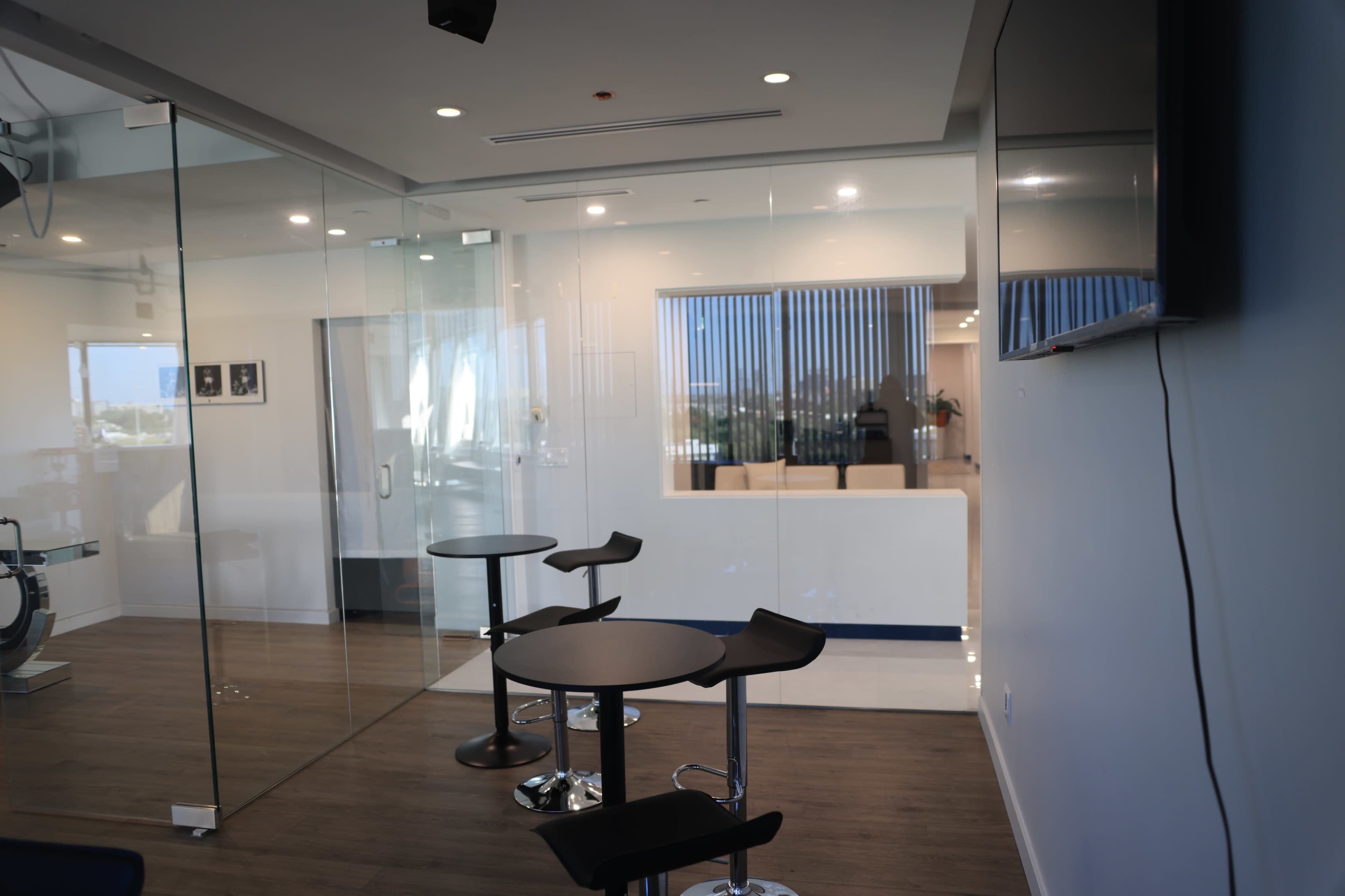 The image shows a modern office space with glass walls, featuring two small round tables and a seating area visible through the glass.