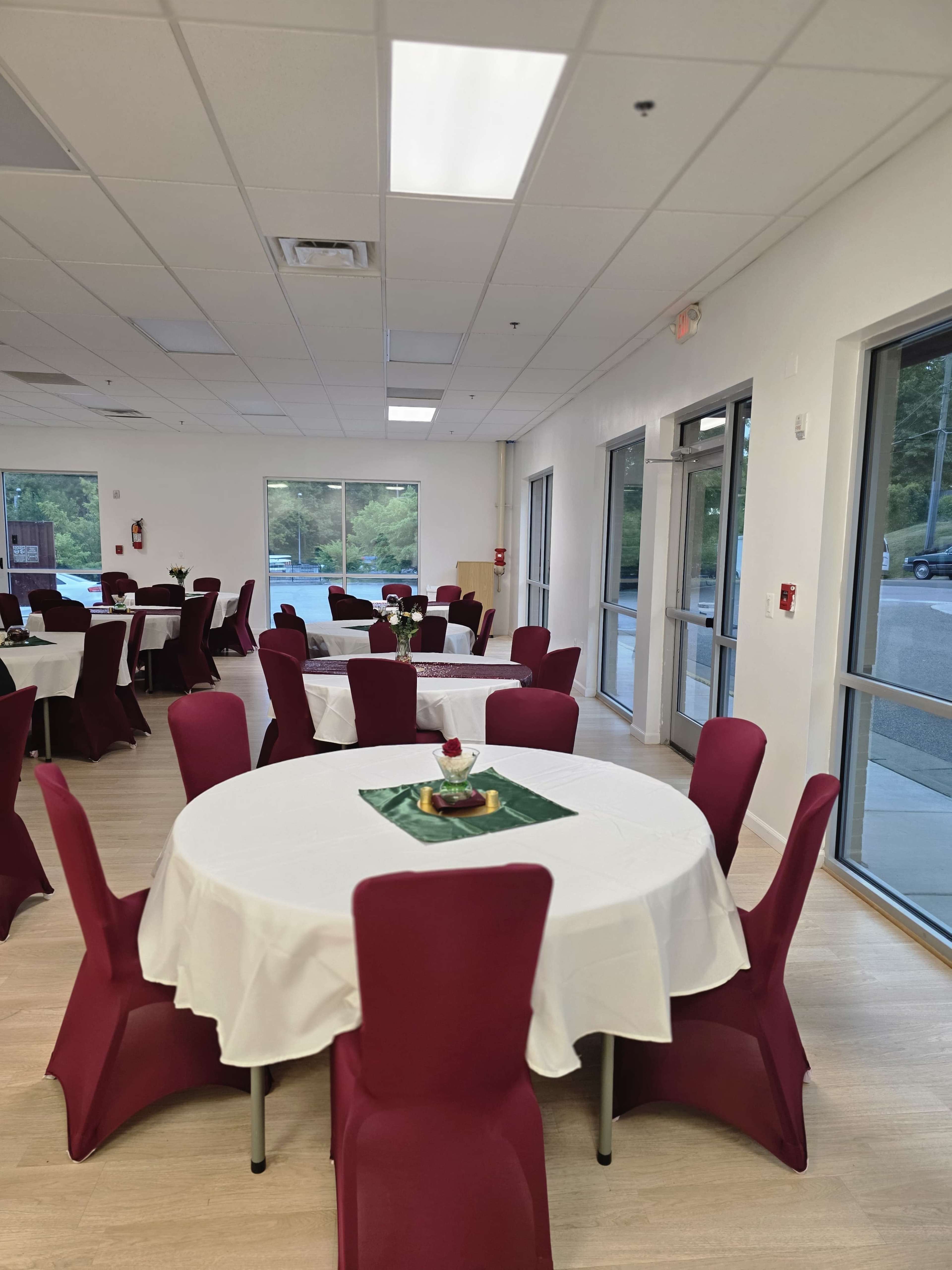 The image shows a spacious dining area with several round tables covered in white tablecloths and surrounded by burgundy chairs.