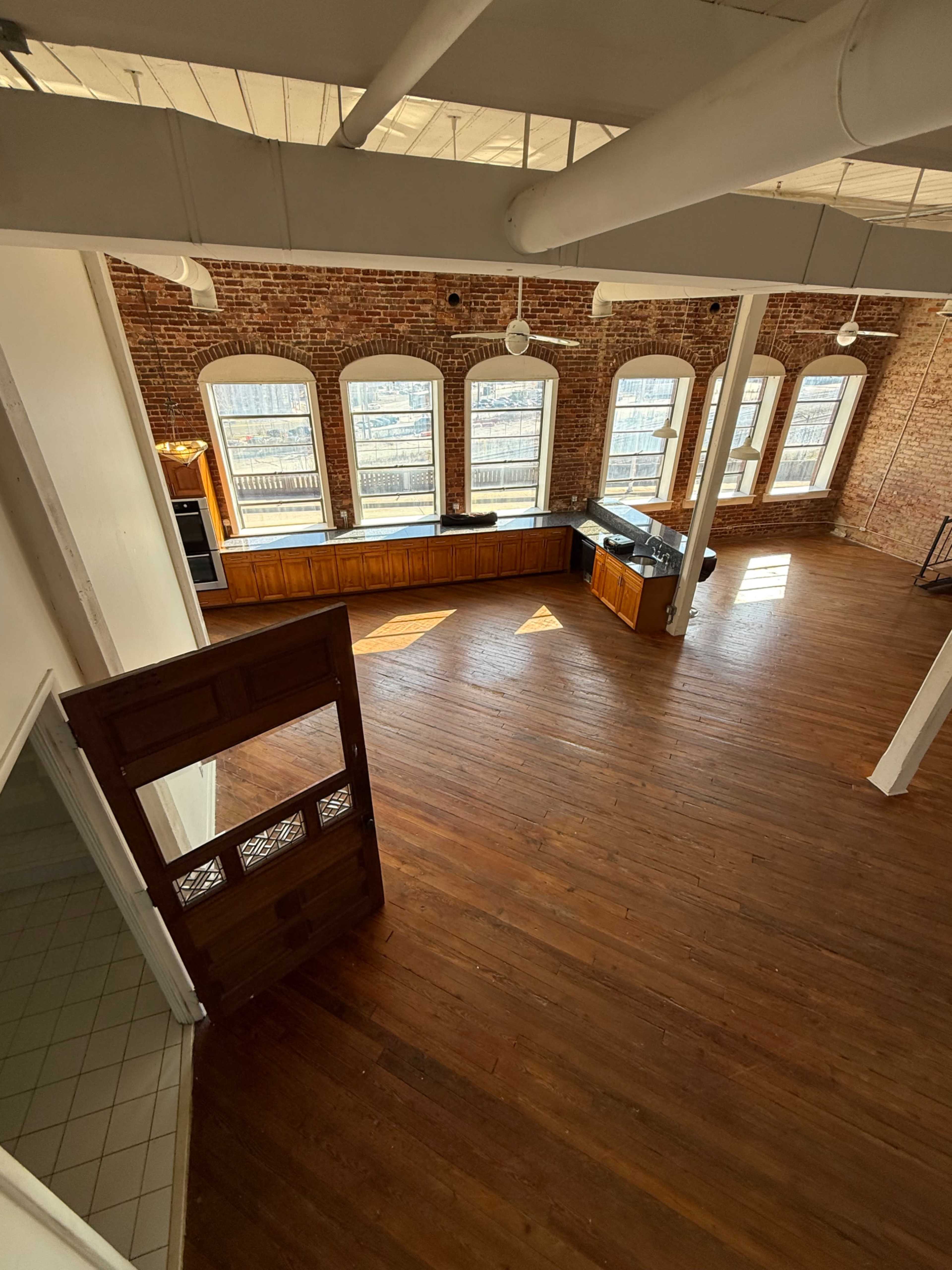 The image shows a spacious loft-style interior with high, exposed brick walls, large windows, and wooden flooring, featuring a kitchen area along one side.