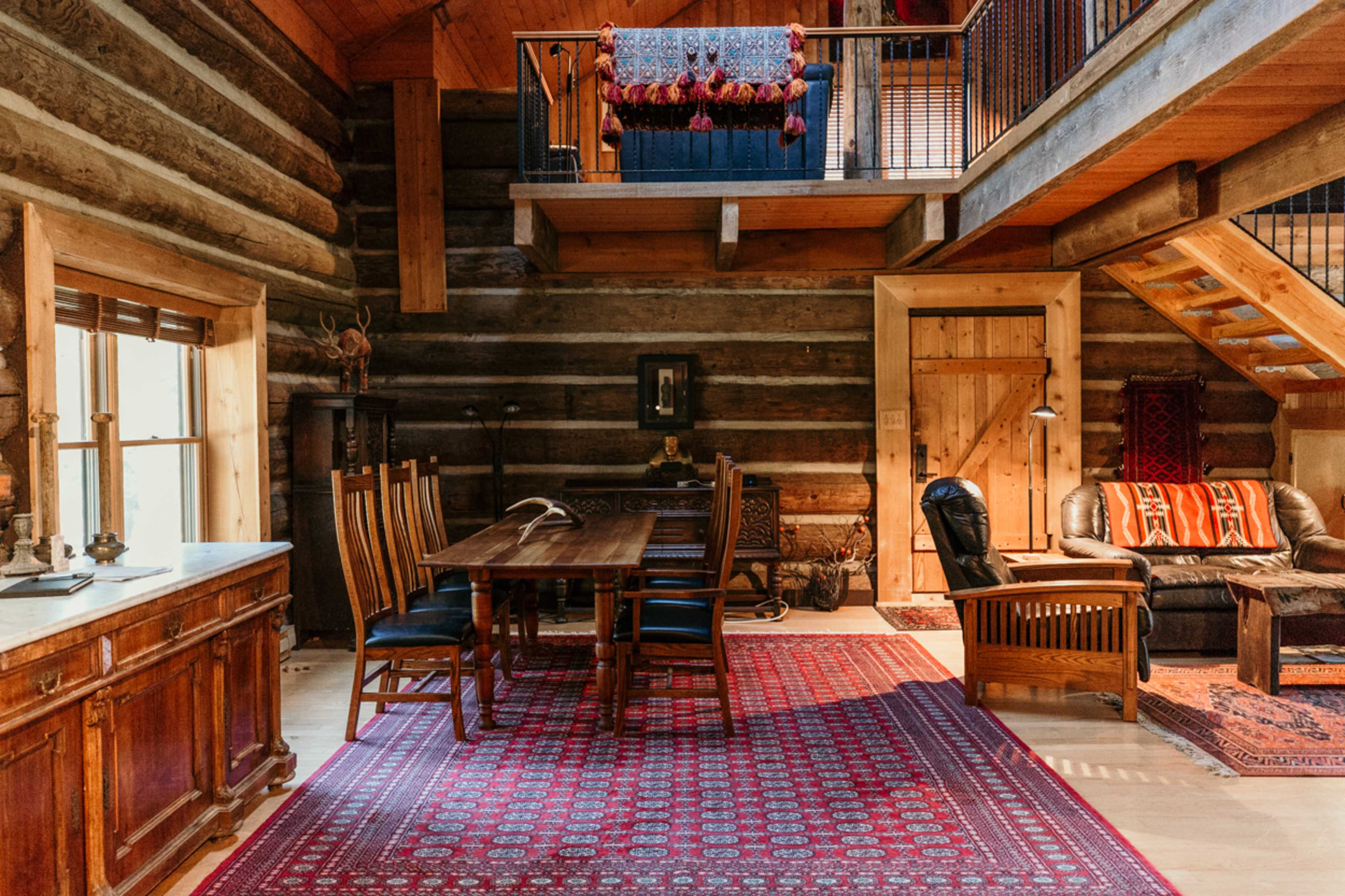 The interior of a rustic log cabin features a dining table with chairs, a staircase leading to a loft, a cozy seating area, and wooden decor elements.