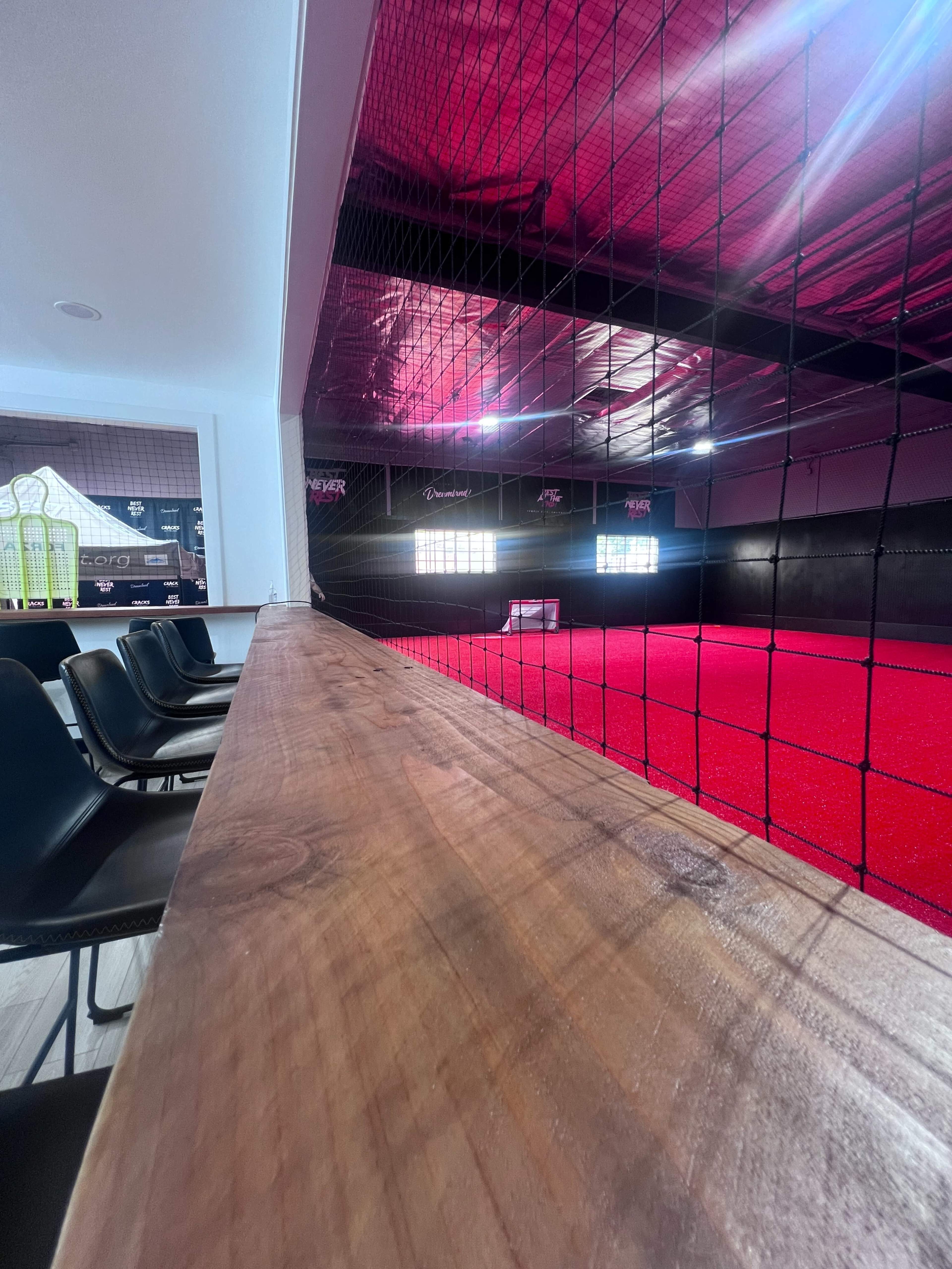 The image shows a modern indoor sports facility with a carpeted playing area, surrounded by a net and featuring wooden seating along the side.