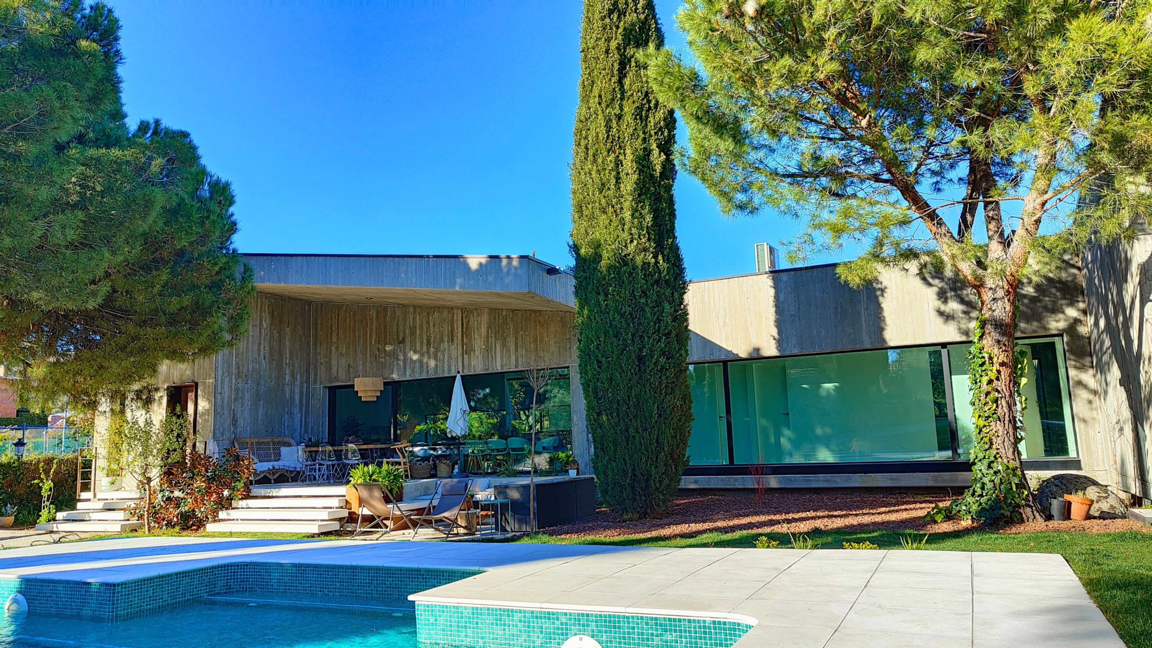 The image shows a modern house with a concrete exterior, large windows, and a swimming pool surrounded by greenery.