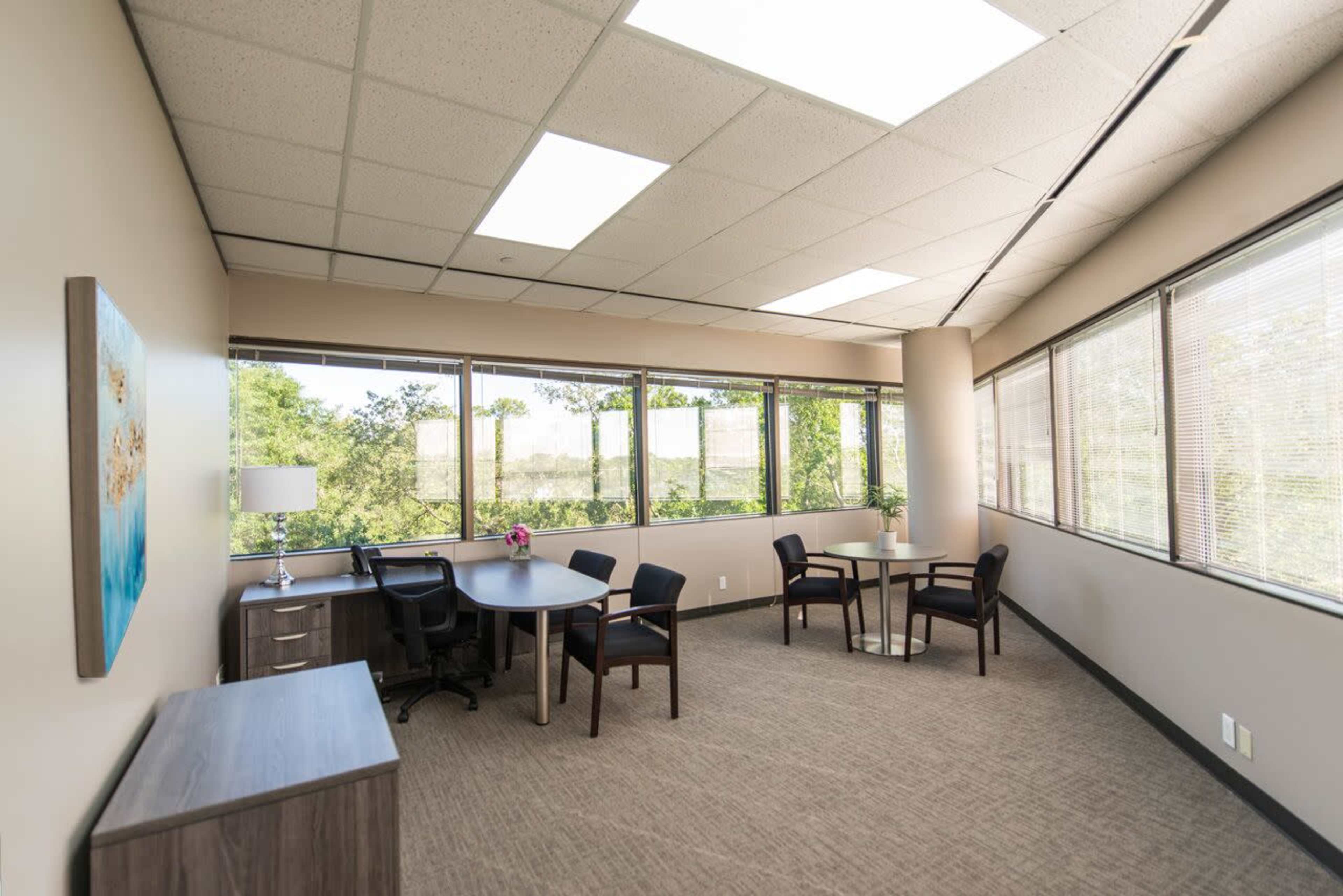 A modern office space with a round table, four chairs, and large windows allowing natural light to enter.