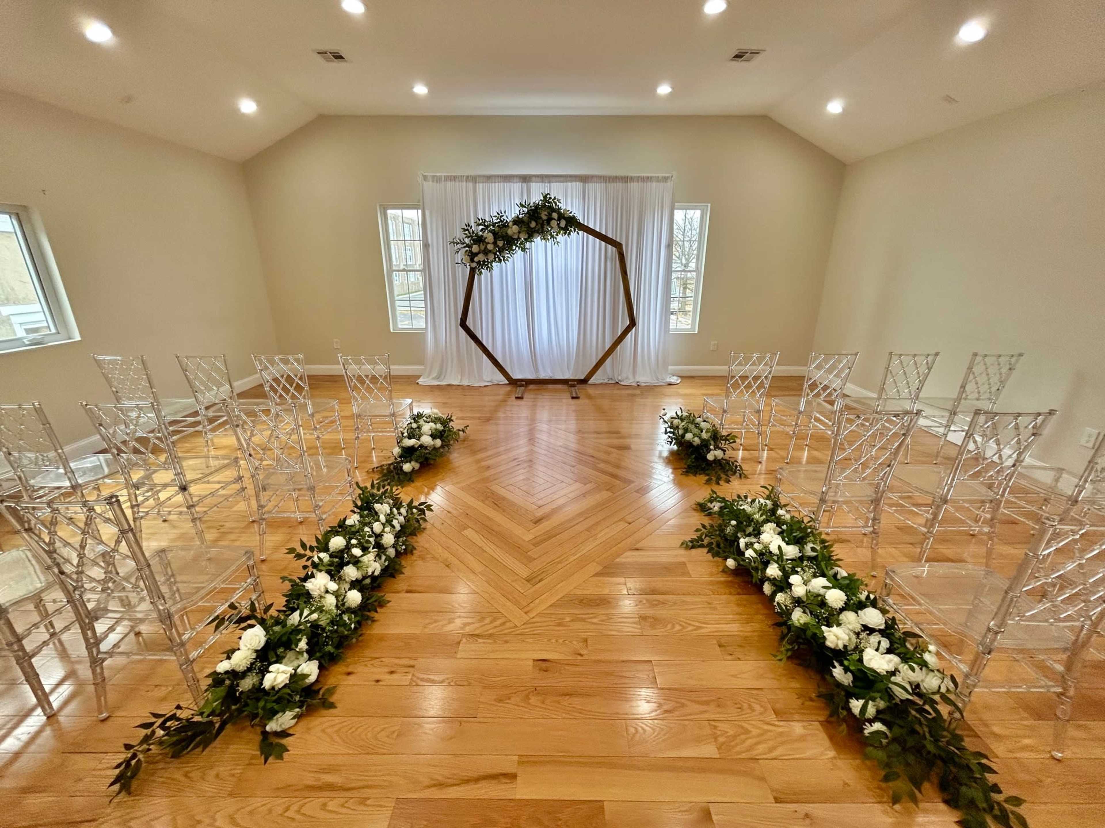 A wedding ceremony setup features transparent chairs arranged in a circular pattern around a wooden arch adorned with greenery, with white flowers lining the aisle.
