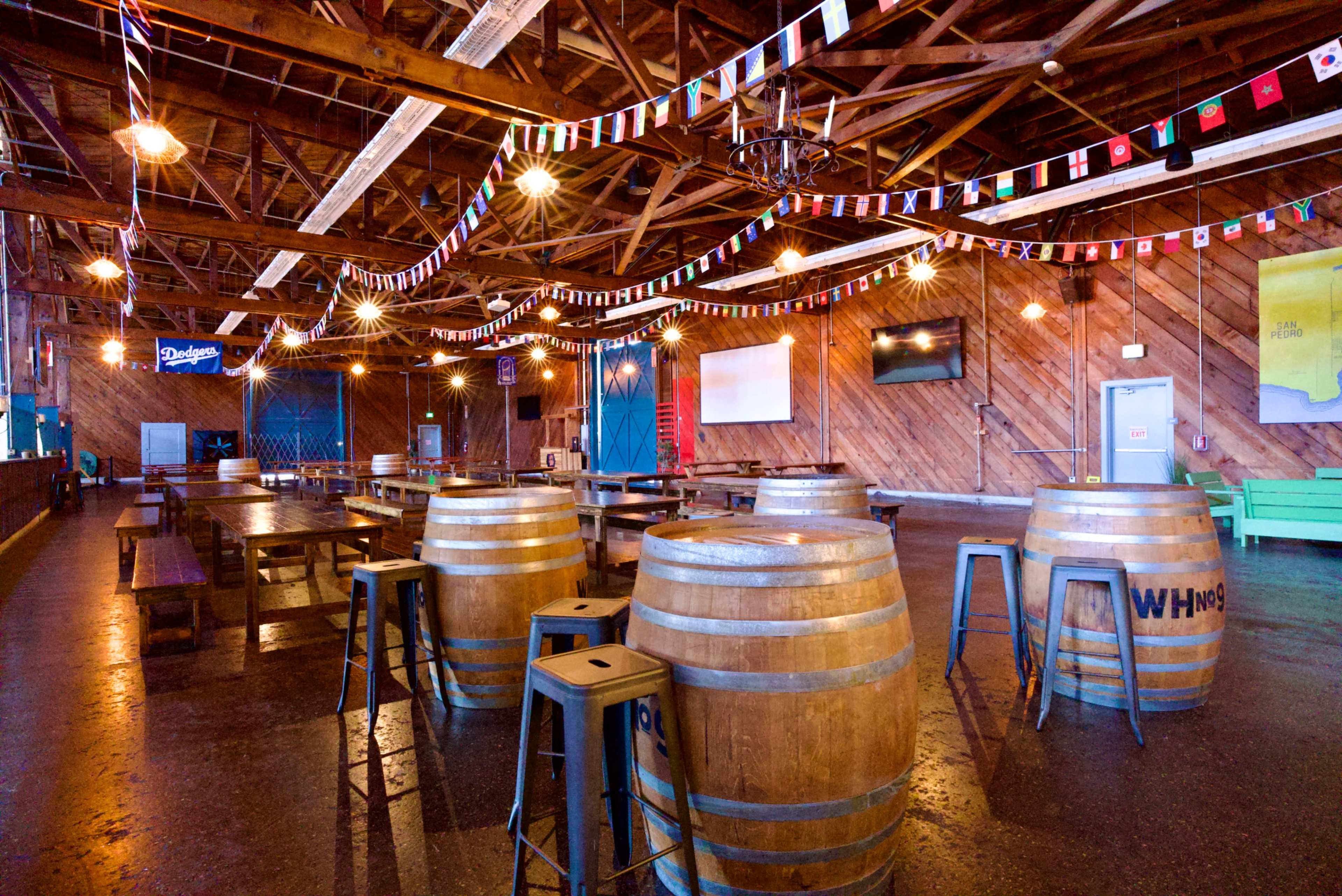 The interior of a rustic venue features wooden barrels, tables, and colorful flags hanging from the ceiling.