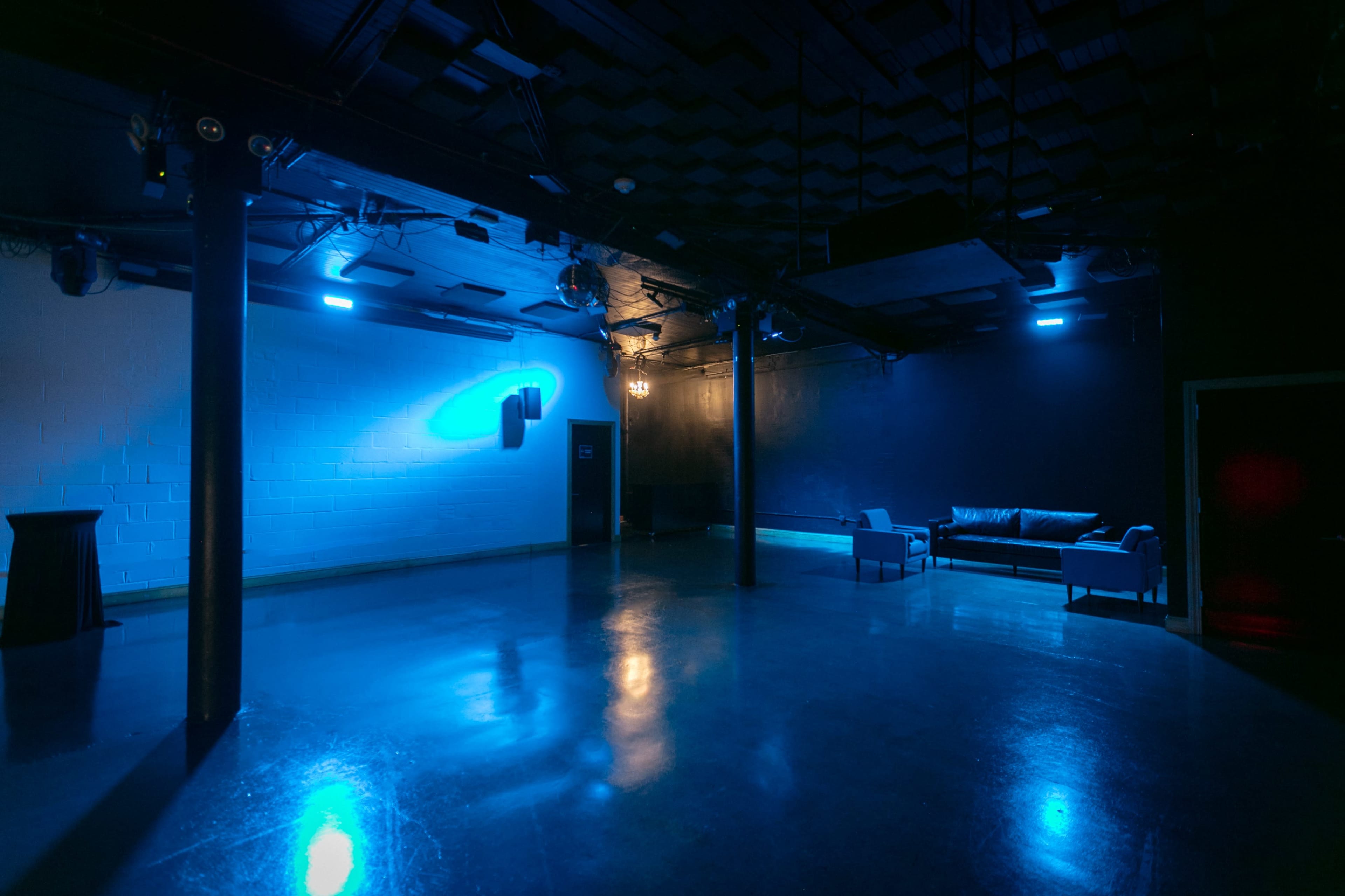 An empty, dimly lit venue with blue lighting, featuring a couch and a tall table in one corner.