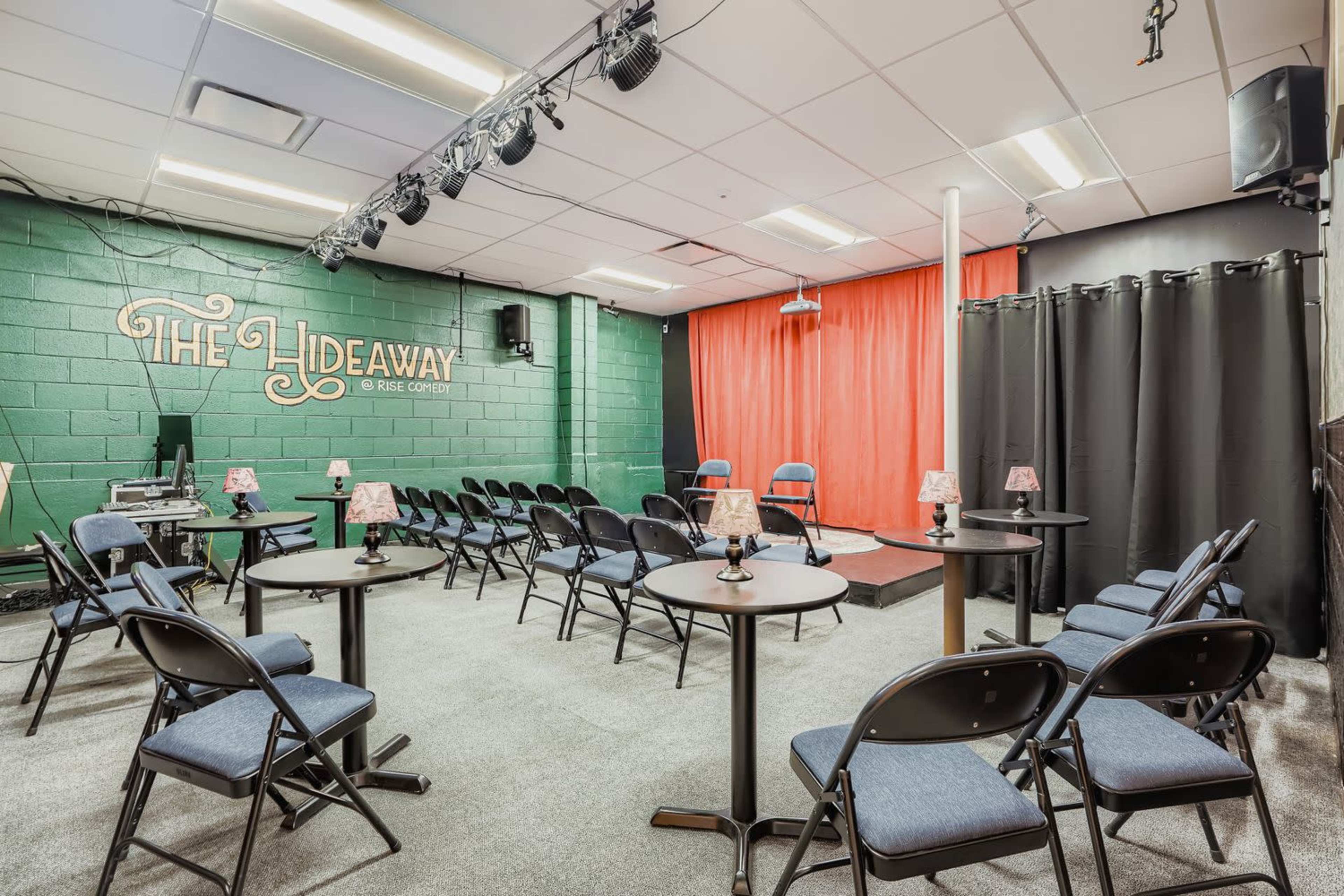 The image shows an intimate performance space with rows of chairs arranged around small tables, featuring a stage with red curtains and soft lighting.