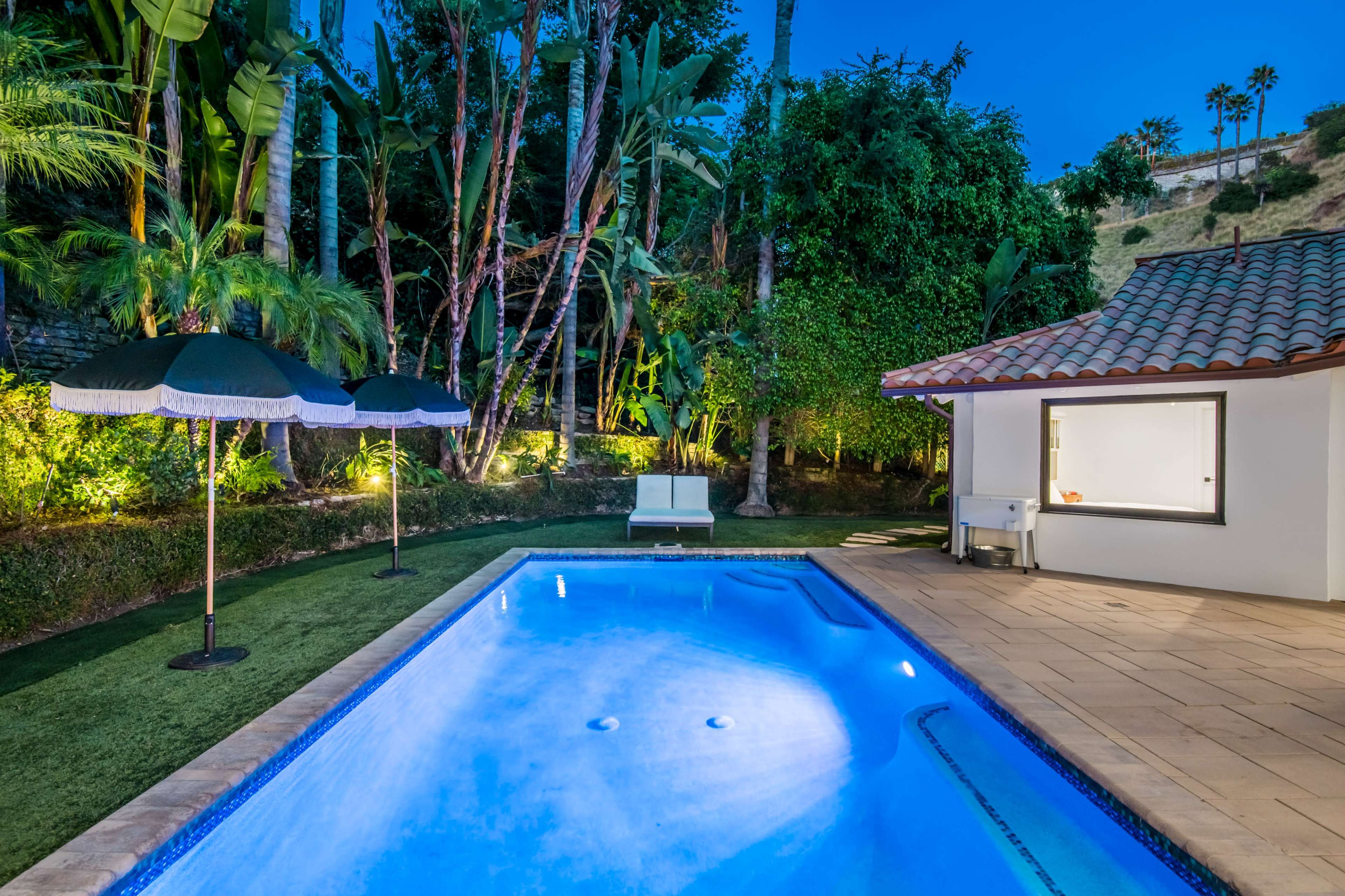 A pool surrounded by tropical plants, with a small lounge area and two black umbrellas positioned nearby.
