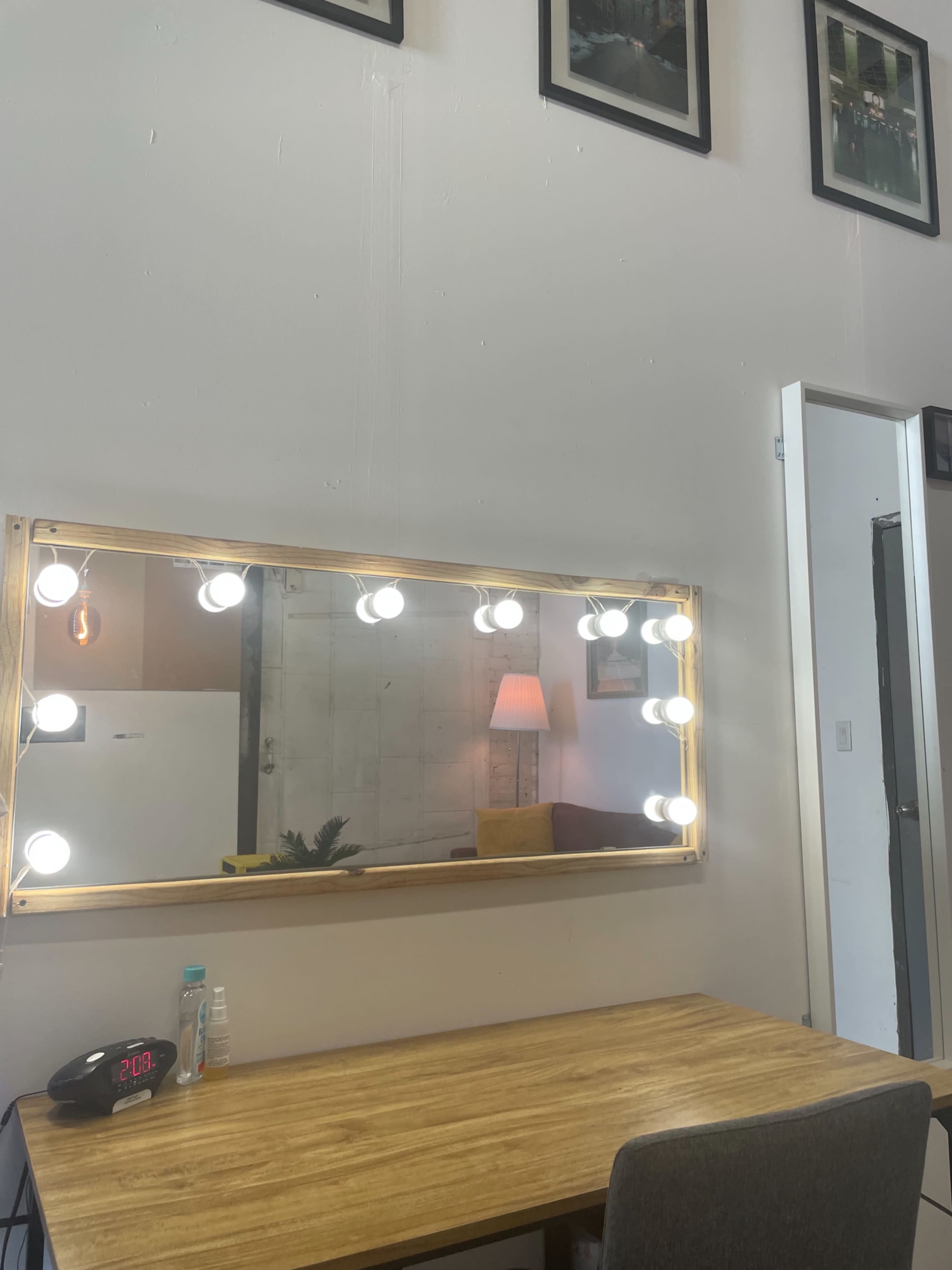 A wooden table in front of a large illuminated mirror, with a desk clock, water bottle, and a lamp visible in the background.