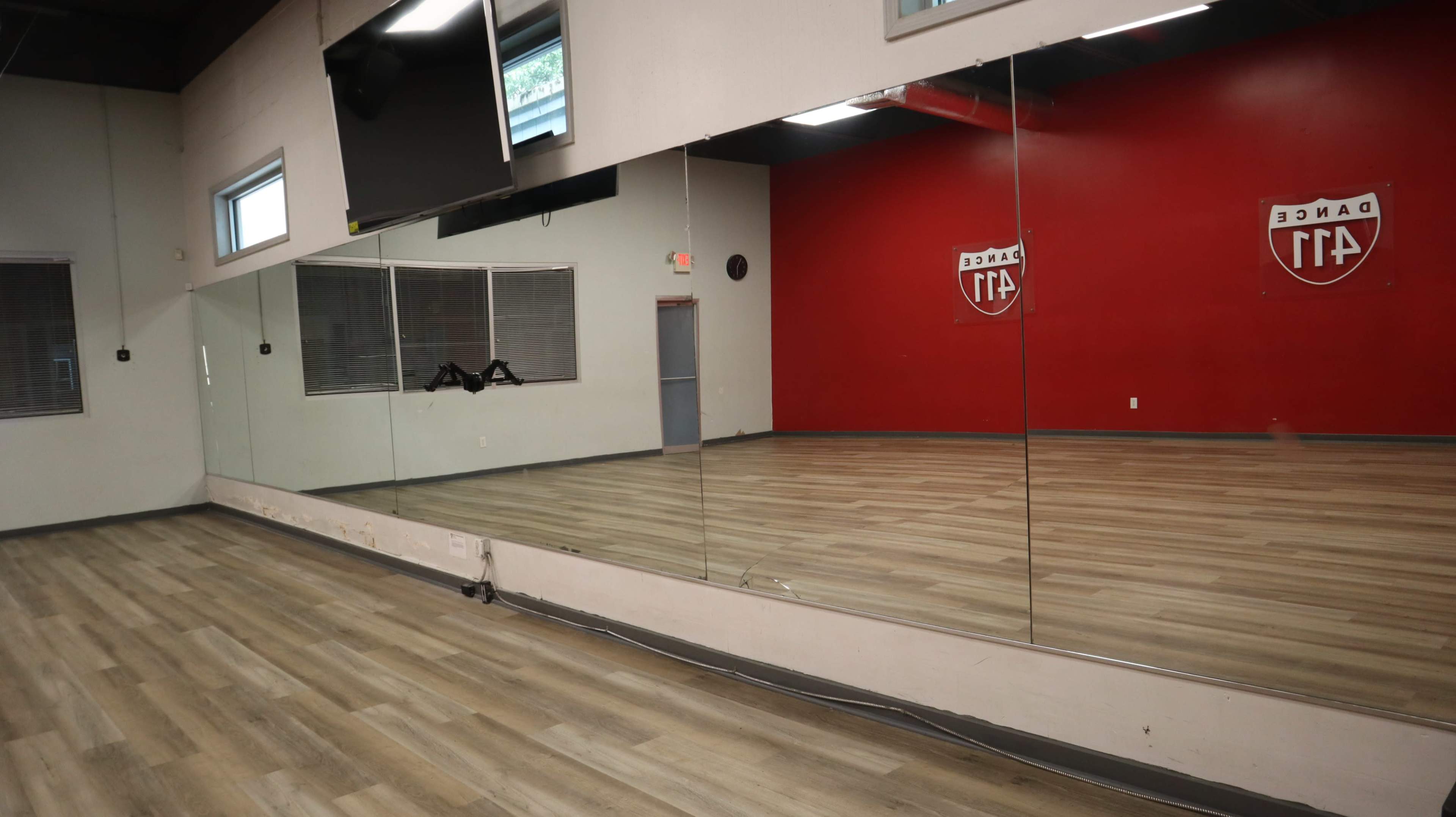 The image shows a dance studio with mirrored walls, wooden flooring, and a red-painted wall featuring a logo.