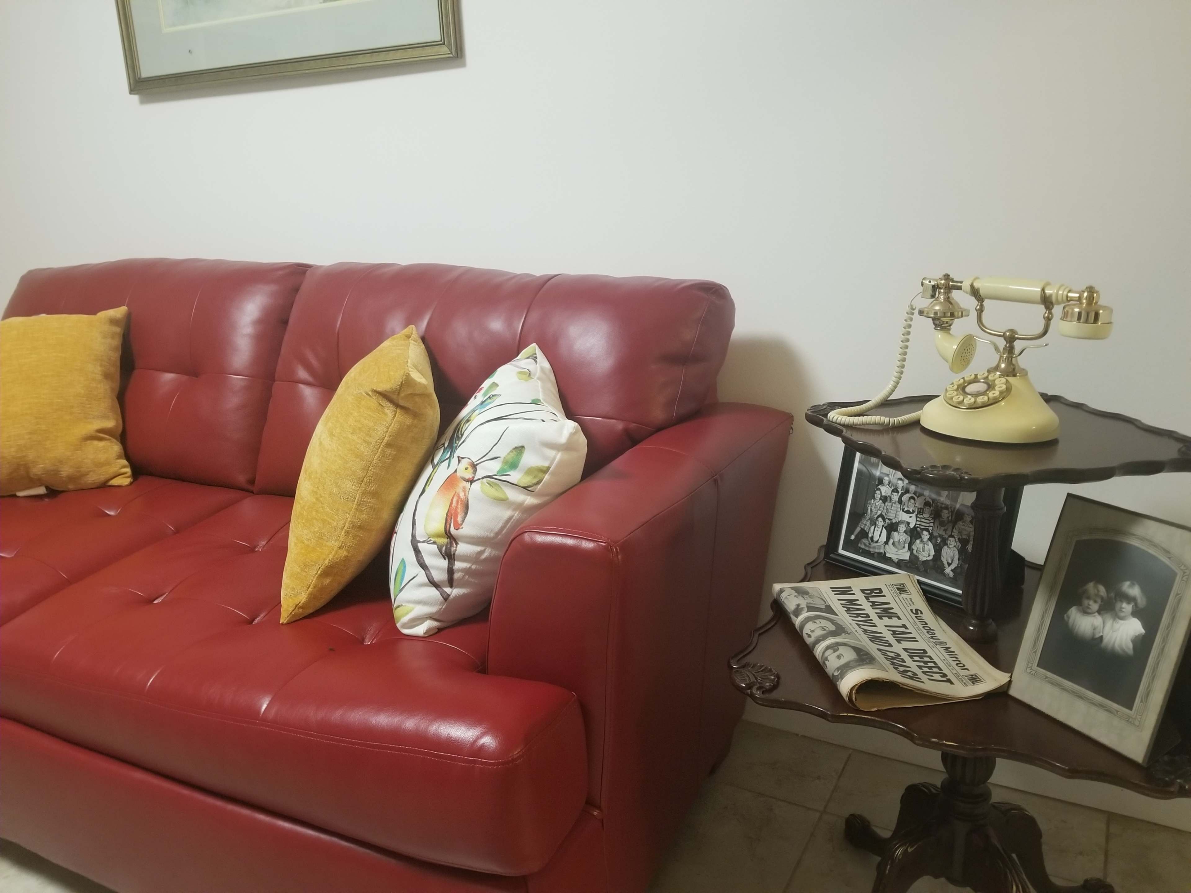 A red leather couch with two pillows, a vintage rotary telephone on a small side table, and a framed black and white photo beside an old newspaper.