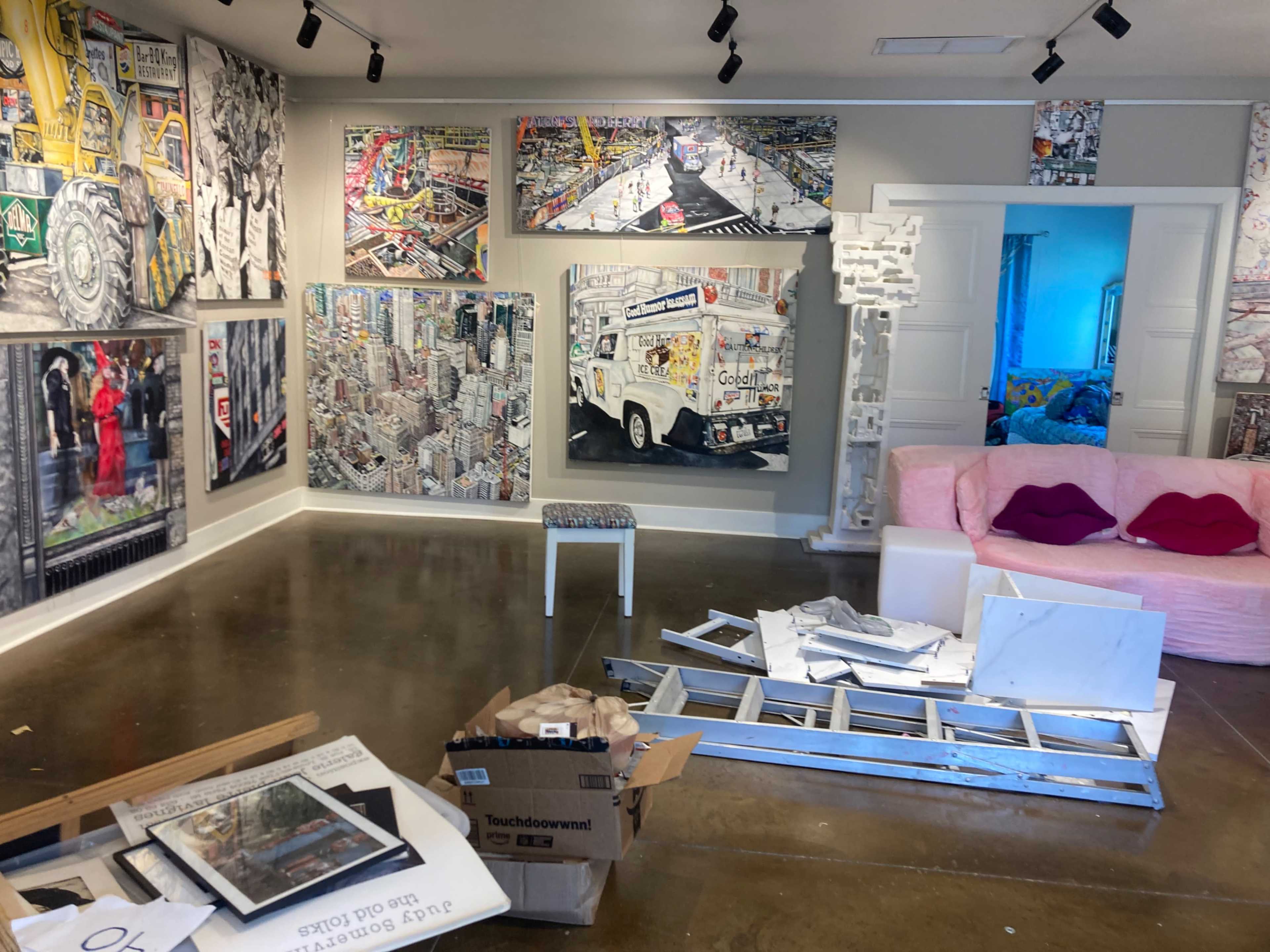 The image shows an art-filled interior space featuring colorful paintings on the walls, discarded materials on the floor, and a pink sofa in the corner.