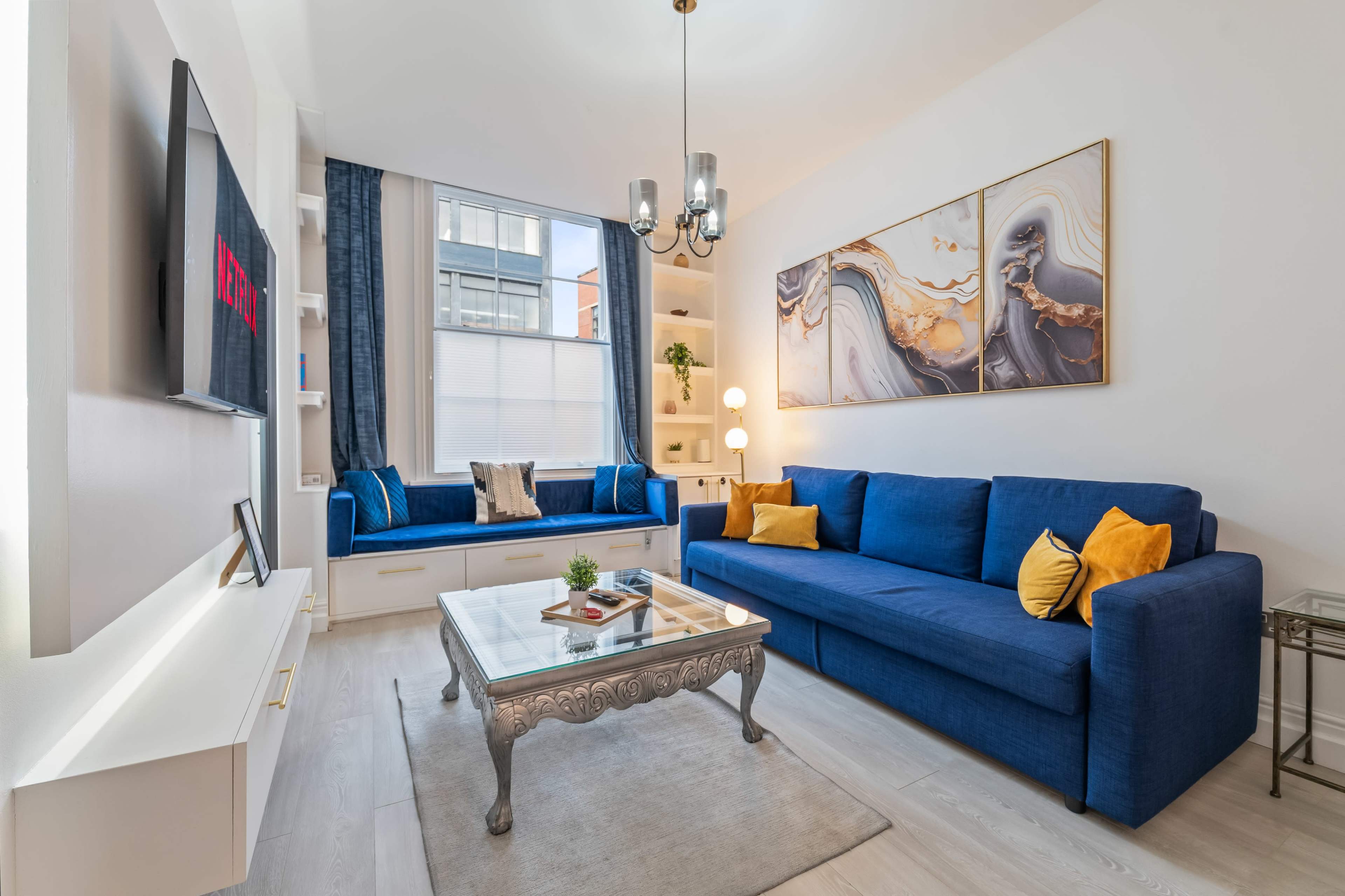 The image shows a modern living room with a blue sofa, a glass coffee table, and two abstract artworks on the wall.