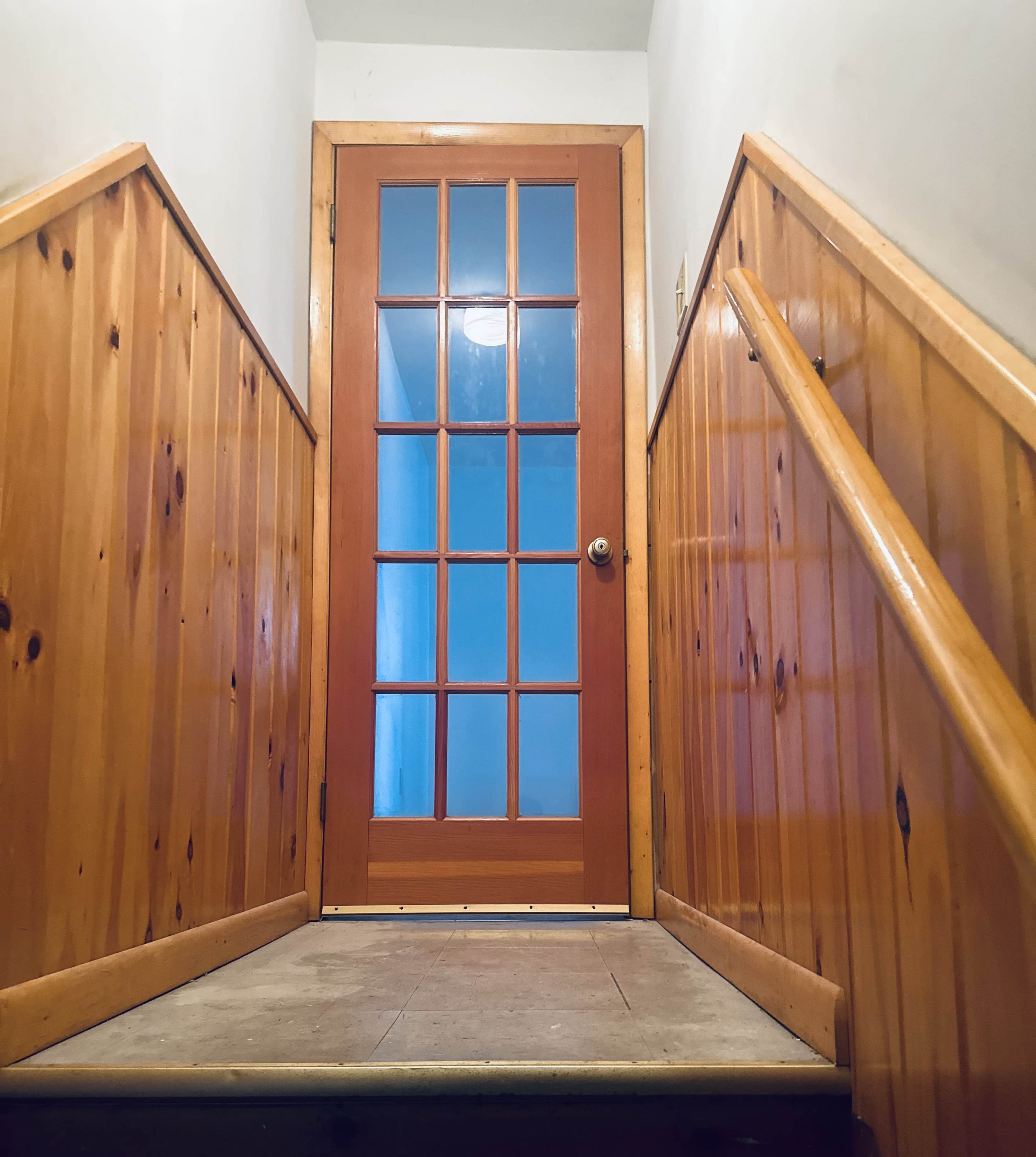 A wooden staircase leads to a glass-paneled door at the top, framed by wooden paneling on the walls.