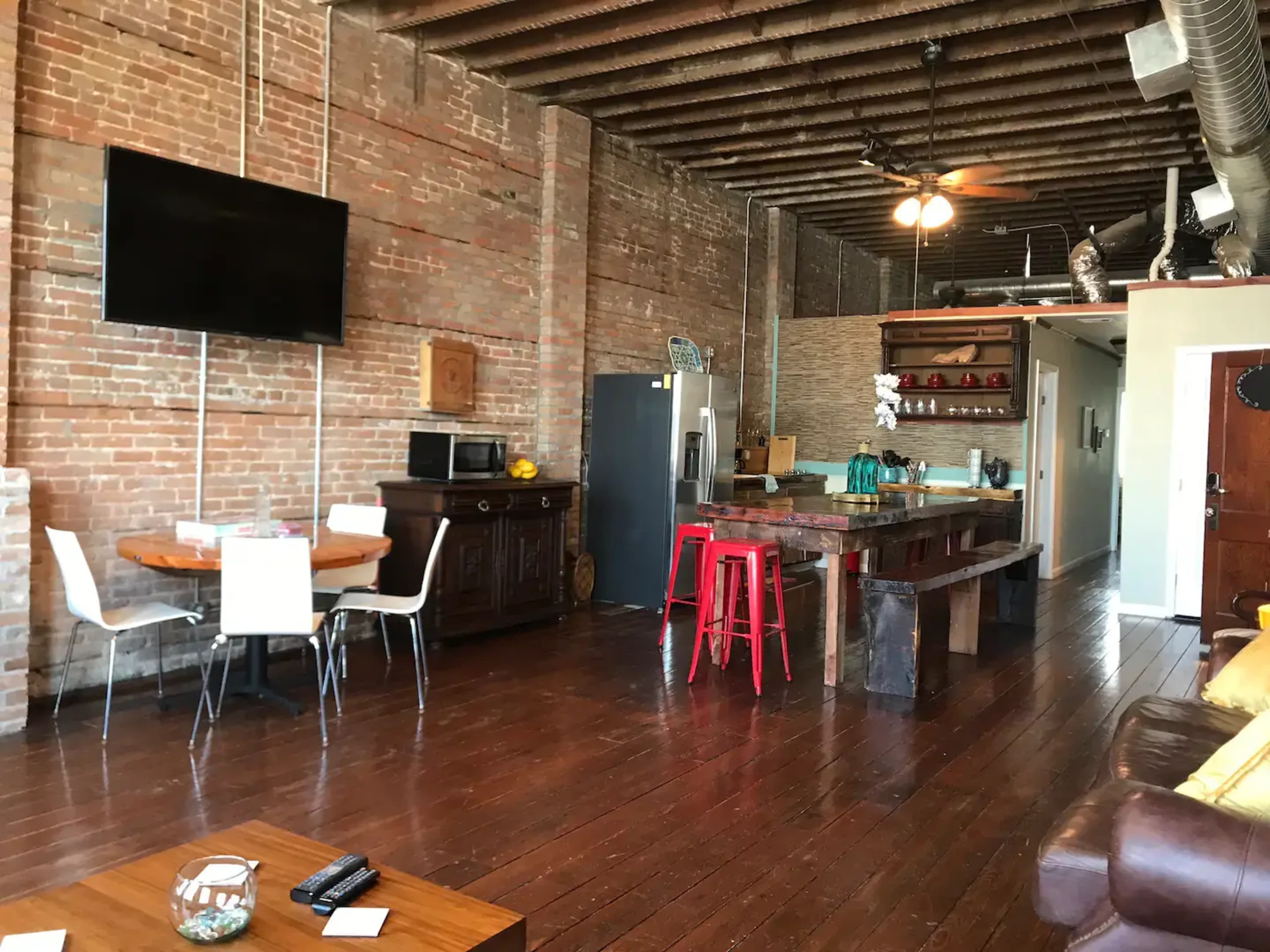 A spacious kitchen and dining area features exposed brick walls, wooden floors, and a large island with red stools.