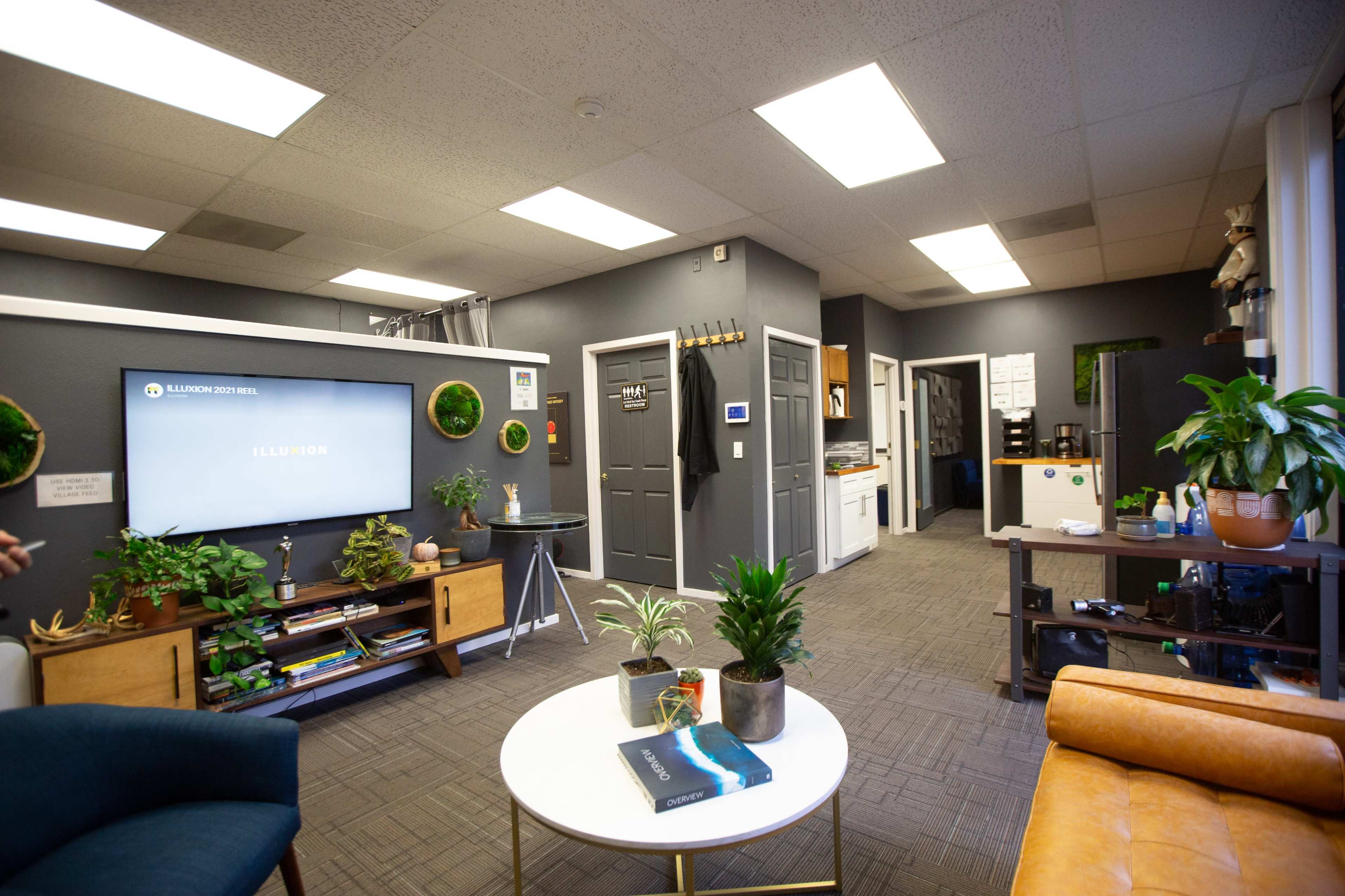 The image shows a modern office space with a seating area, a large screen on the wall displaying "ILLUMION," and various plants and decorative elements throughout the room.