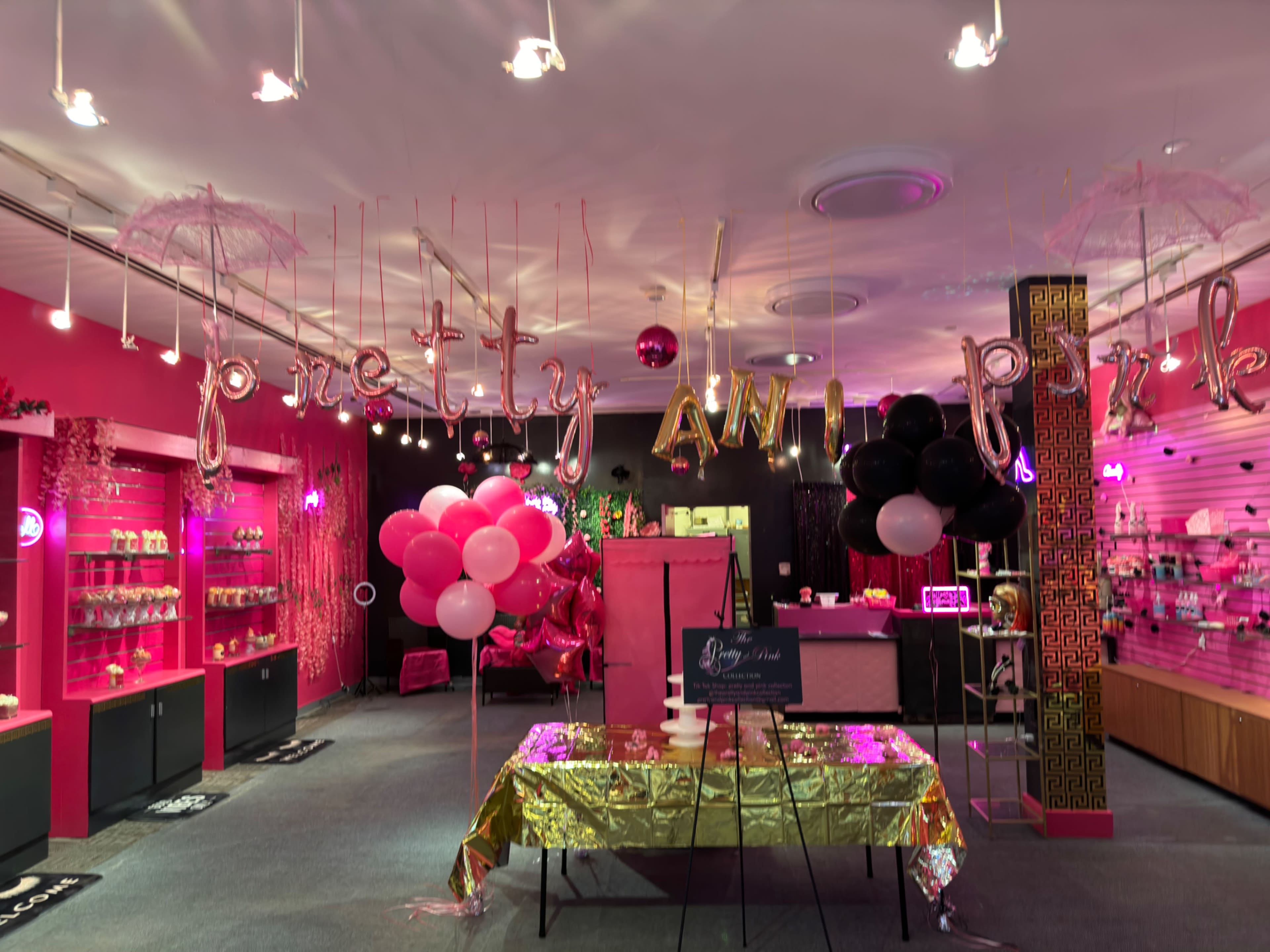 The interior of a vibrant boutique features pink décor, colorful balloons, and a table covered in a gold tablecloth for a celebration.