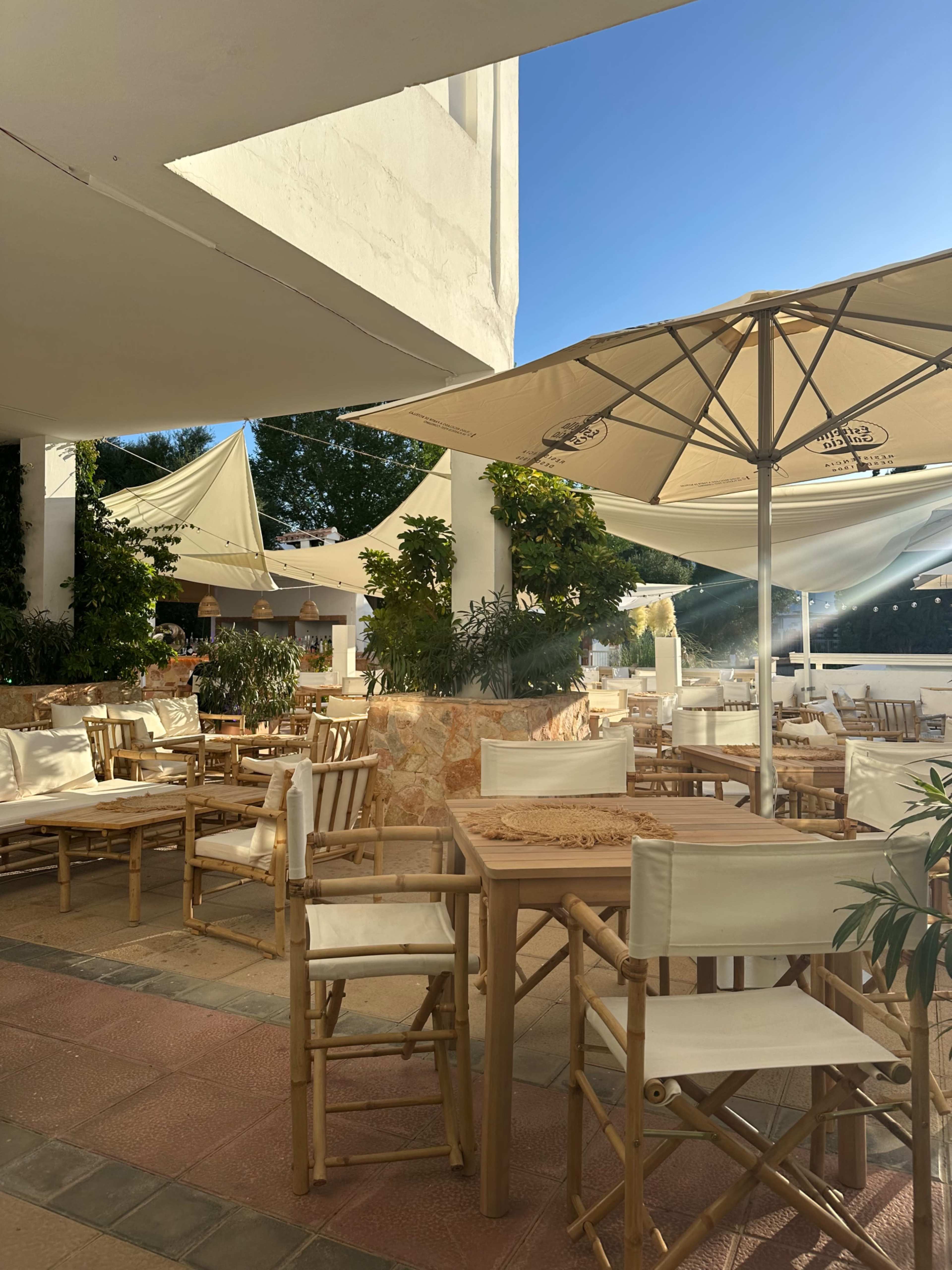 A sunlit outdoor seating area features wooden tables and chairs surrounded by greenery and shaded by large umbrellas.
