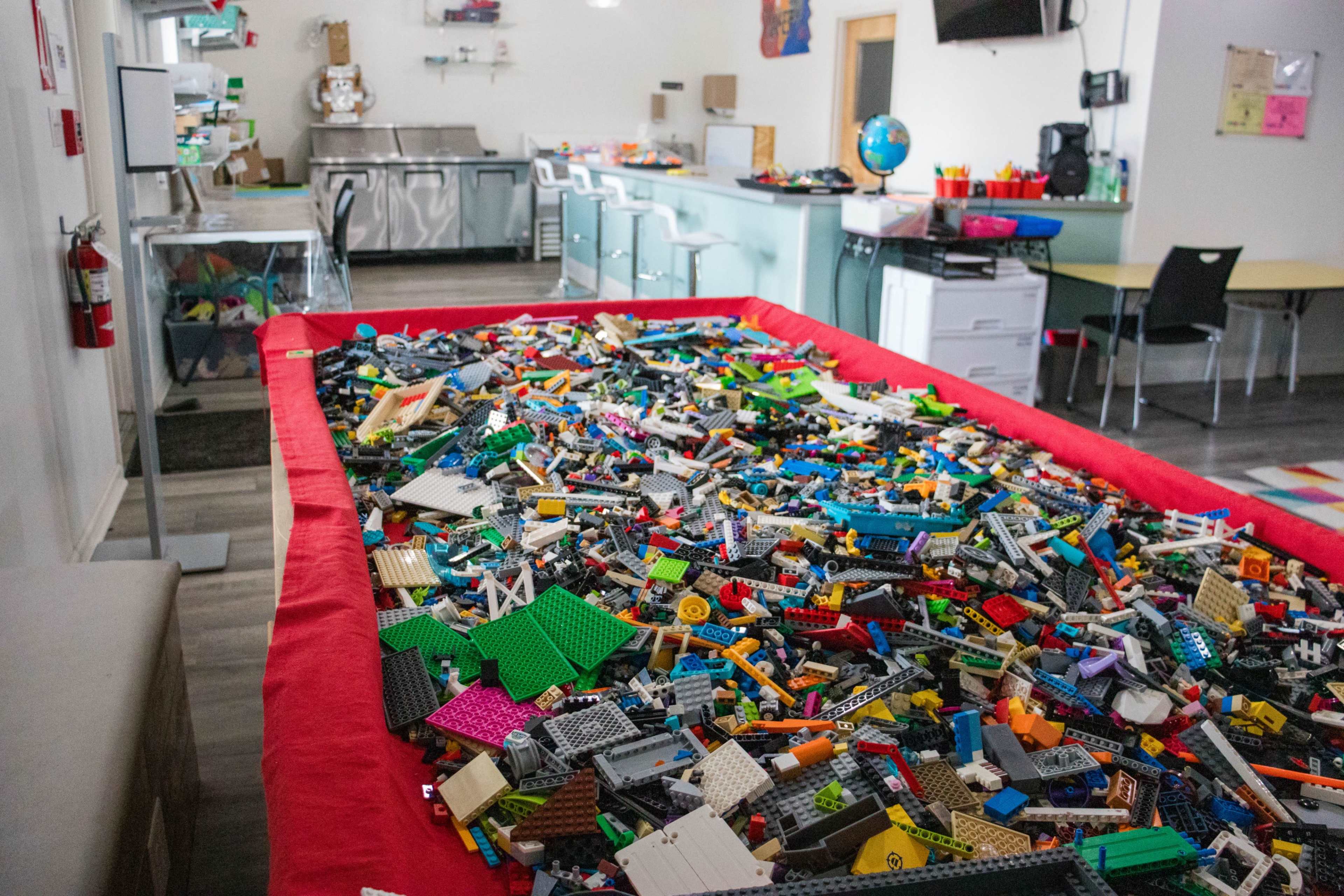A large red table is filled with various colorful Lego pieces in a bright, open room with kitchen and work areas in the background.