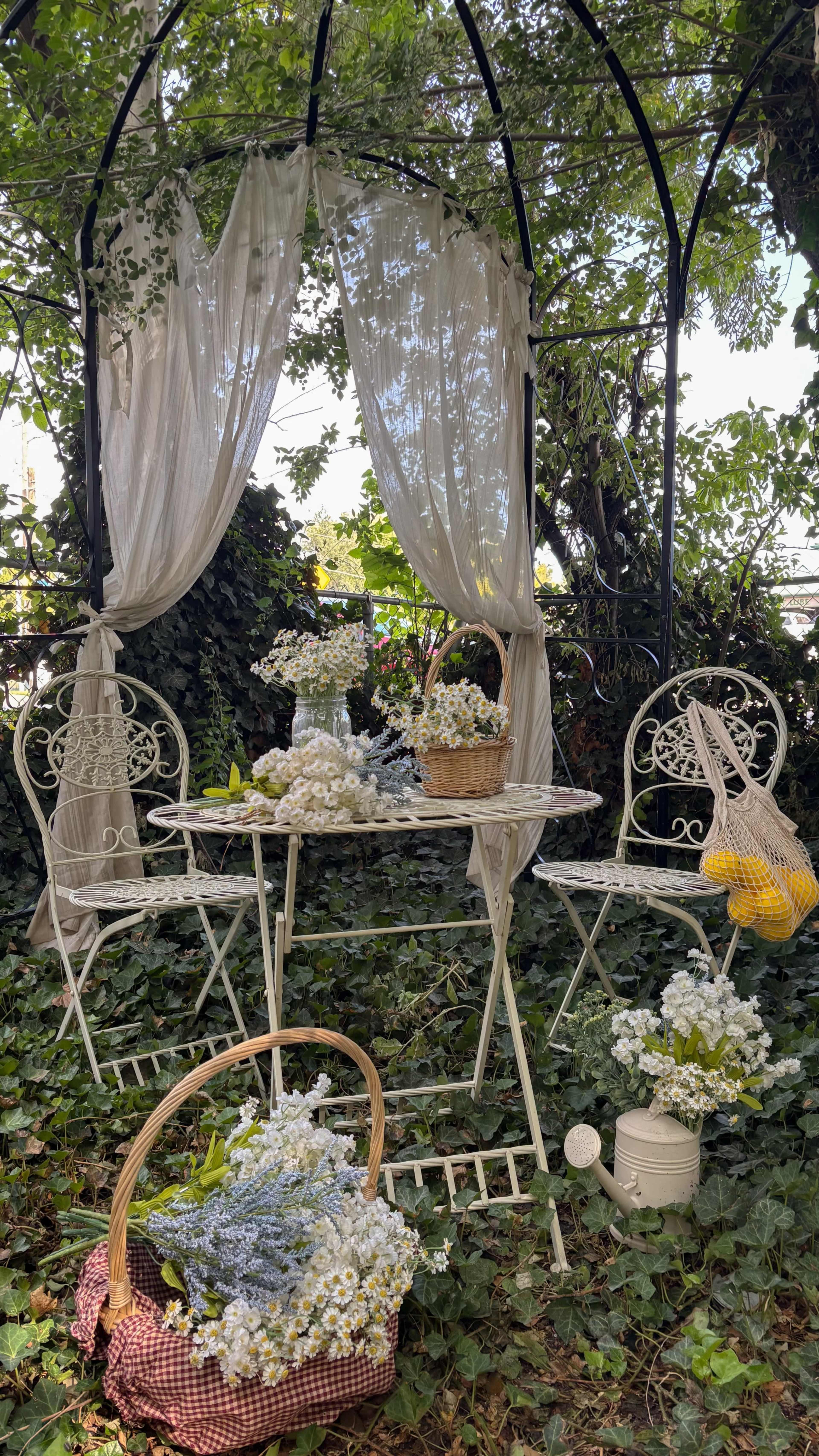 A white, wrought-iron gazebo is adorned with sheer curtains and surrounded by greenery, featuring a table set with baskets of flowers and two chairs.