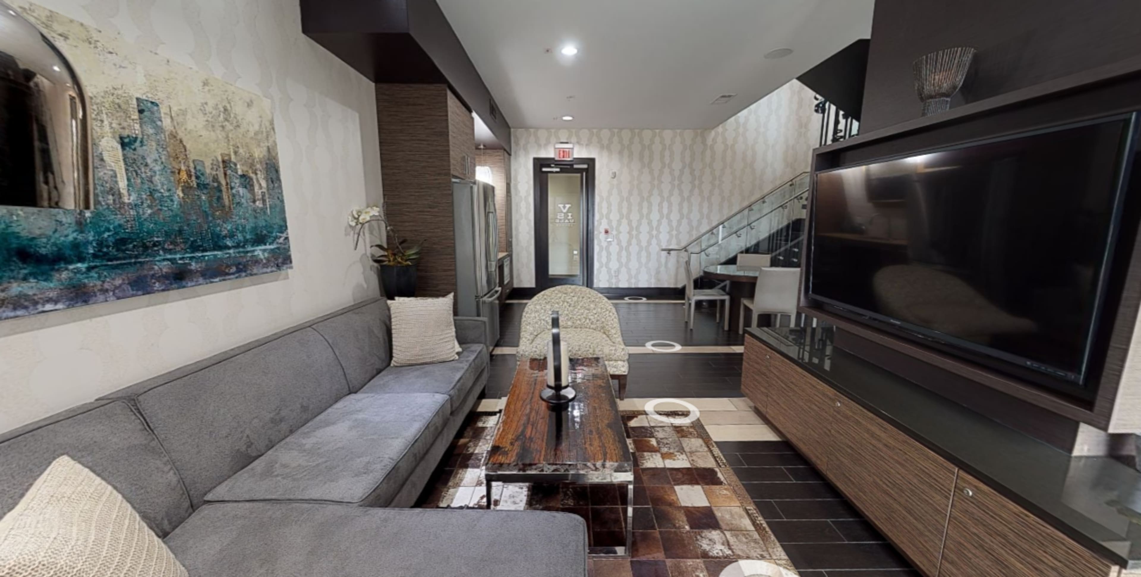 A modern living space with a gray sofa, a wooden coffee table, and a large television positioned against a wall, featuring a decorative painting and a staircase in the background.