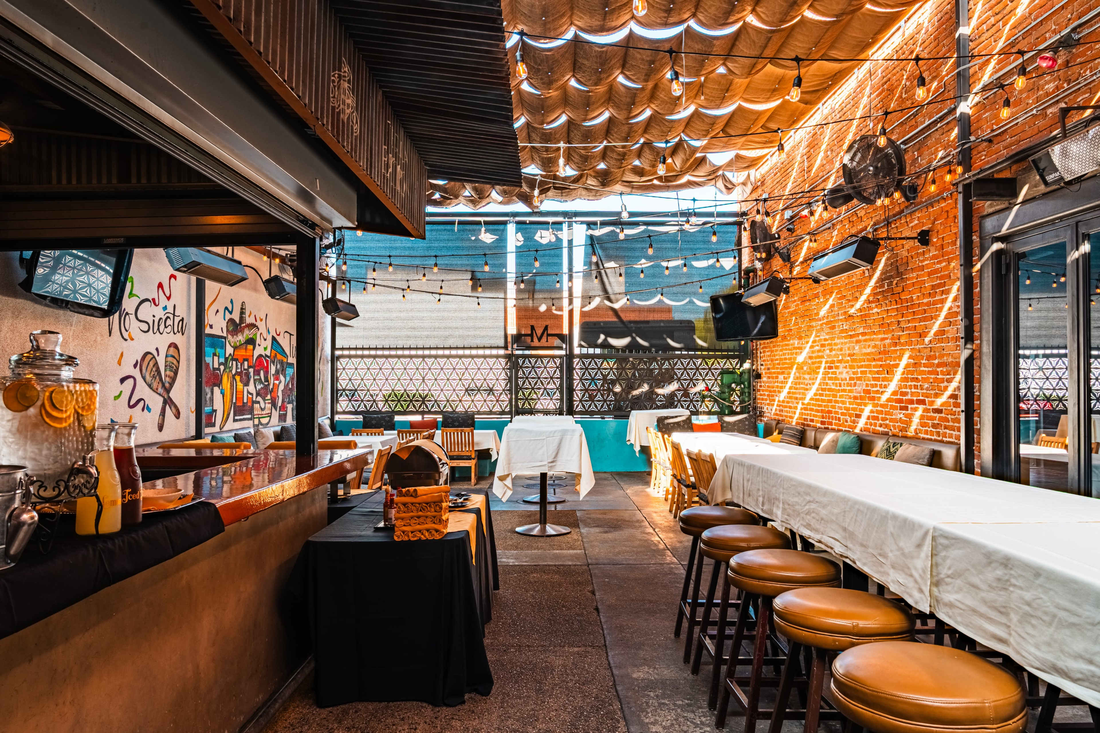 The image shows an empty outdoor dining area of a restaurant with a long table draped in white cloth, wooden stools, and string lights overhead.