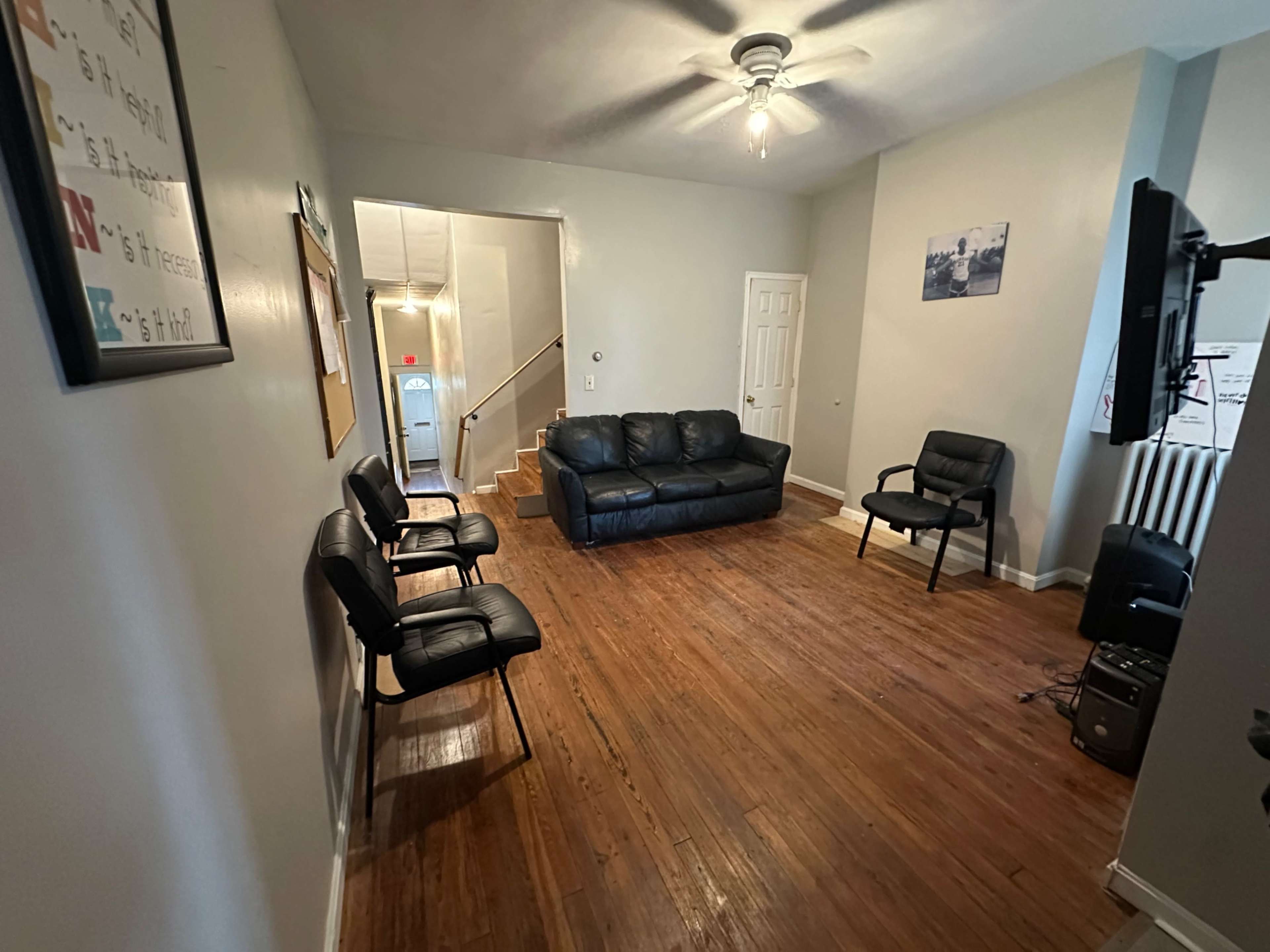 The image shows a living room with a black leather couch, two black chairs, and wooden flooring, leading to a staircase at the back.