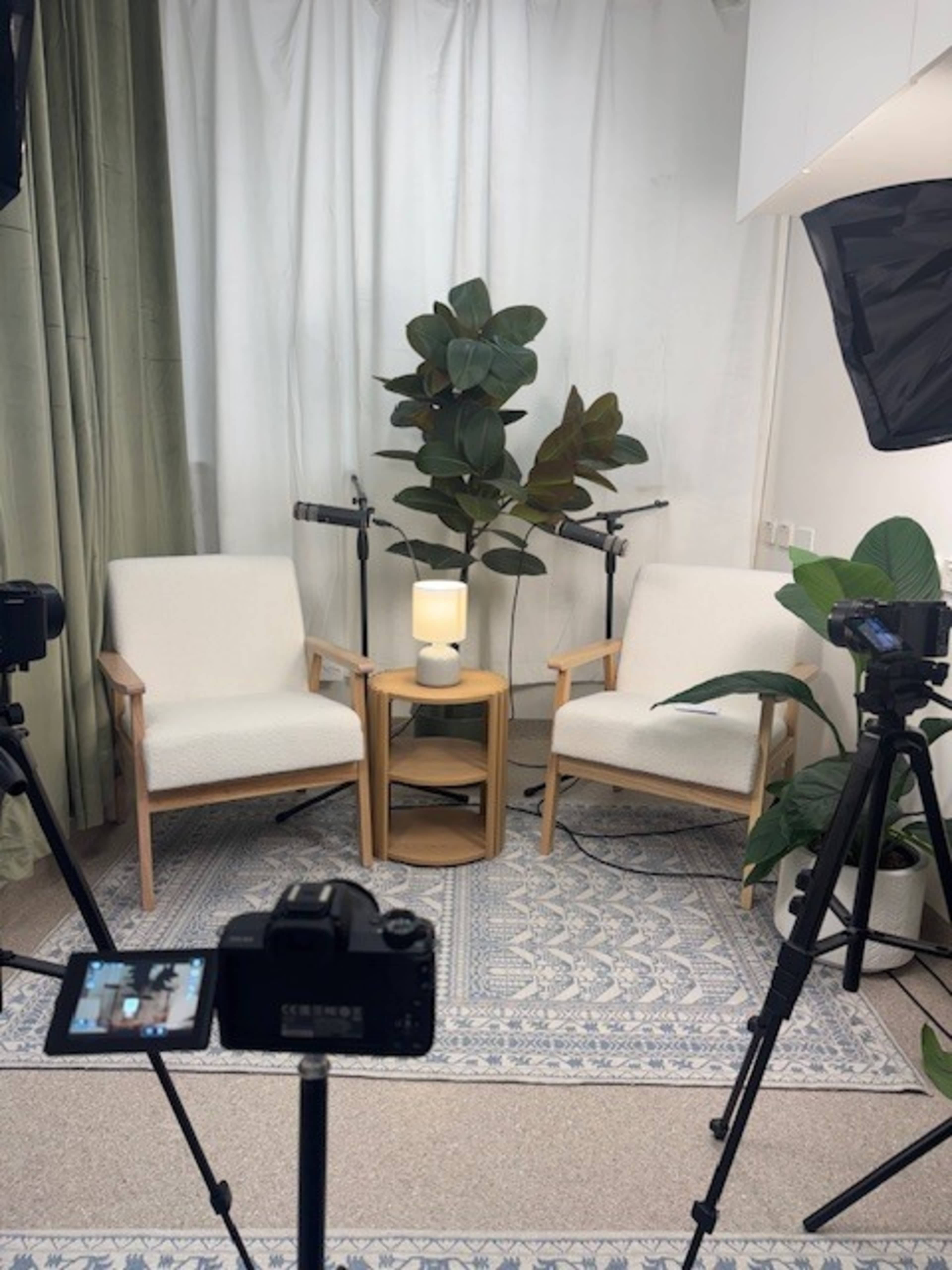 The image shows a cozy interview setup featuring two white chairs, a small wooden table with a lamp, and decorative plants, all arranged in a softly lit room.