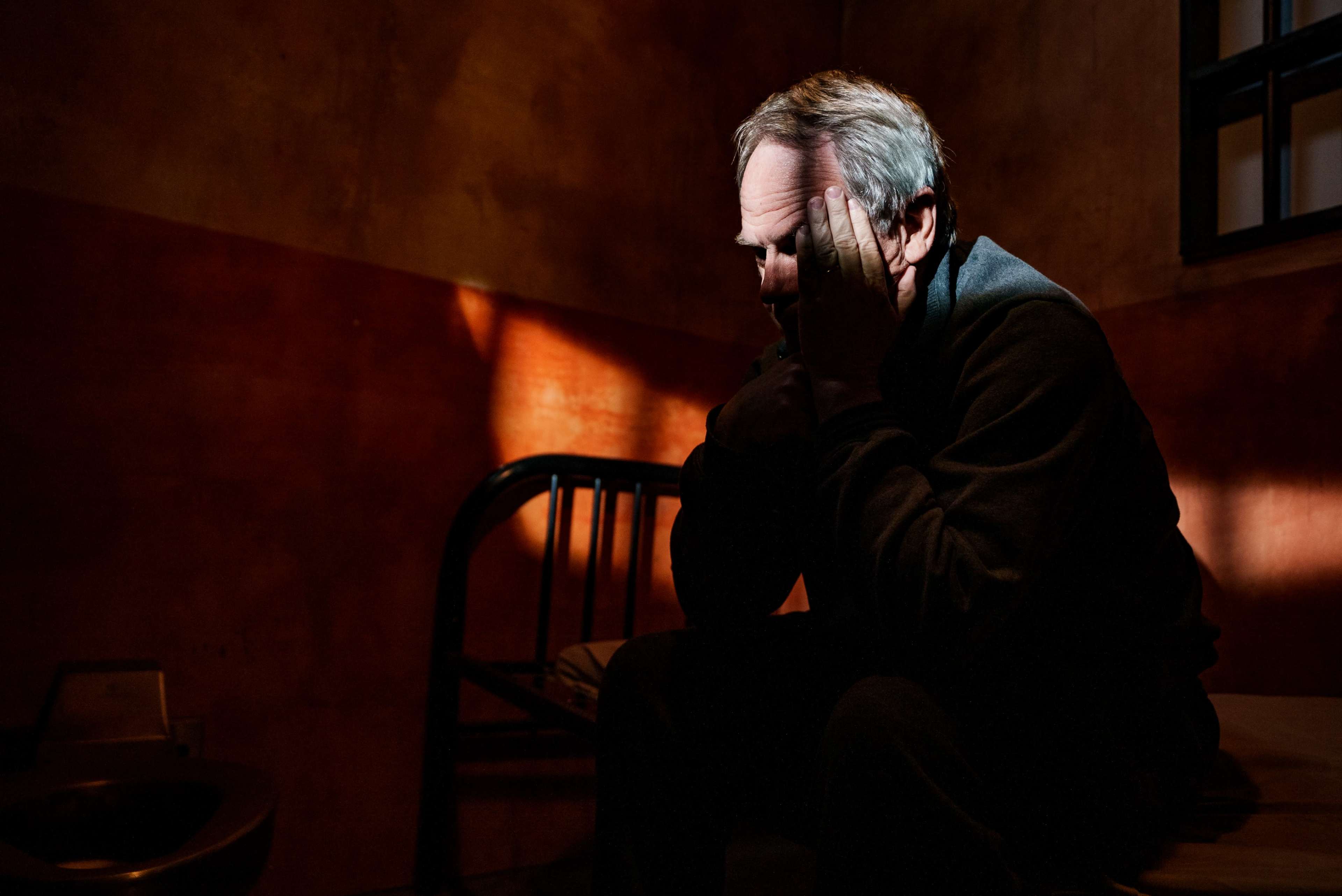 A man sits on the edge of a bed in a dimly lit room, holding his head in his hands with shadows cast on the walls.