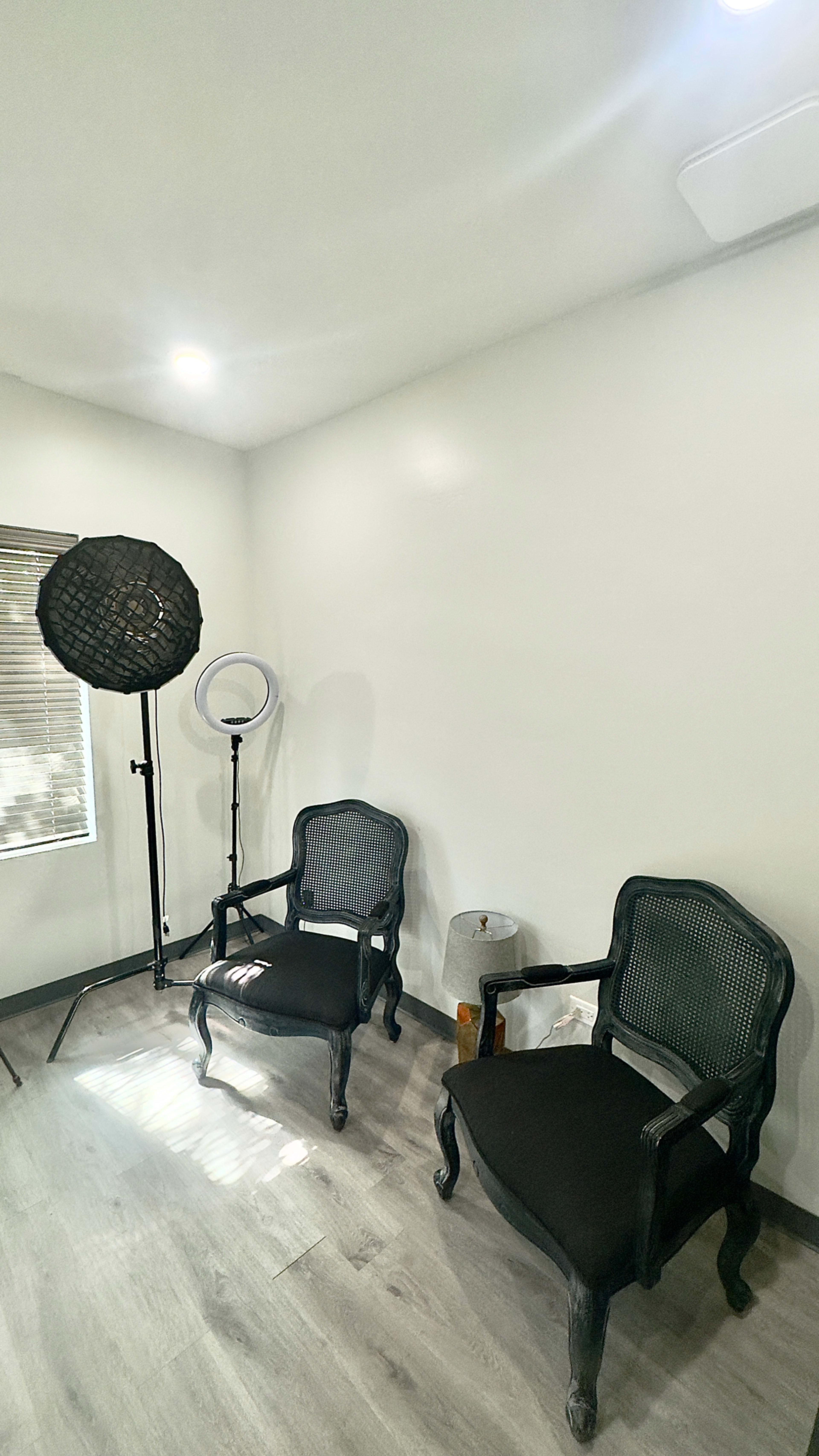 The image shows a small, well-lit room with two black chairs, a round light ring, and a light stand, alongside a decorative lamp and a white bag.