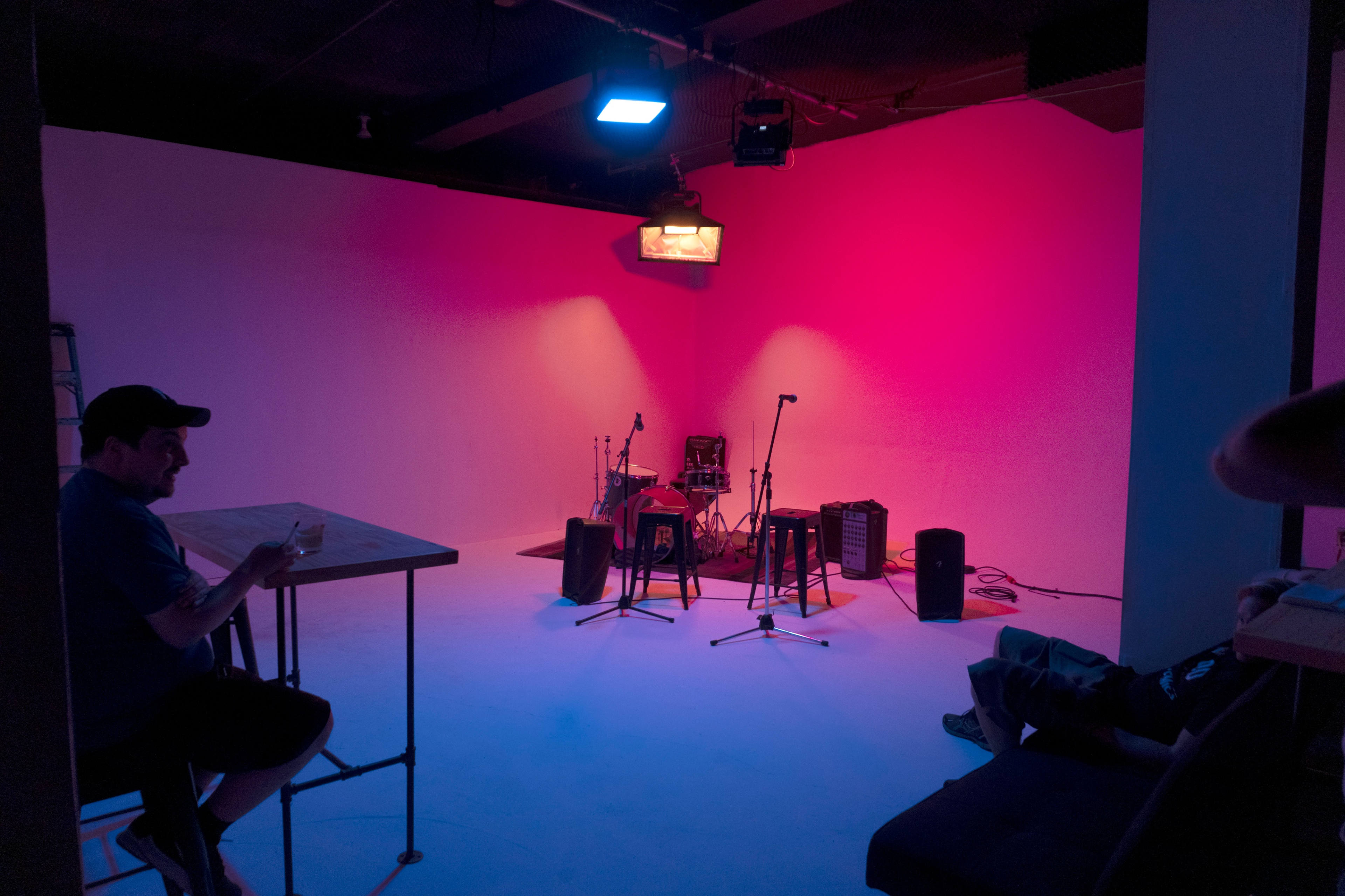 A dimly lit studio features a small stage setup with musical instruments and two chairs, while a man sits at a table.