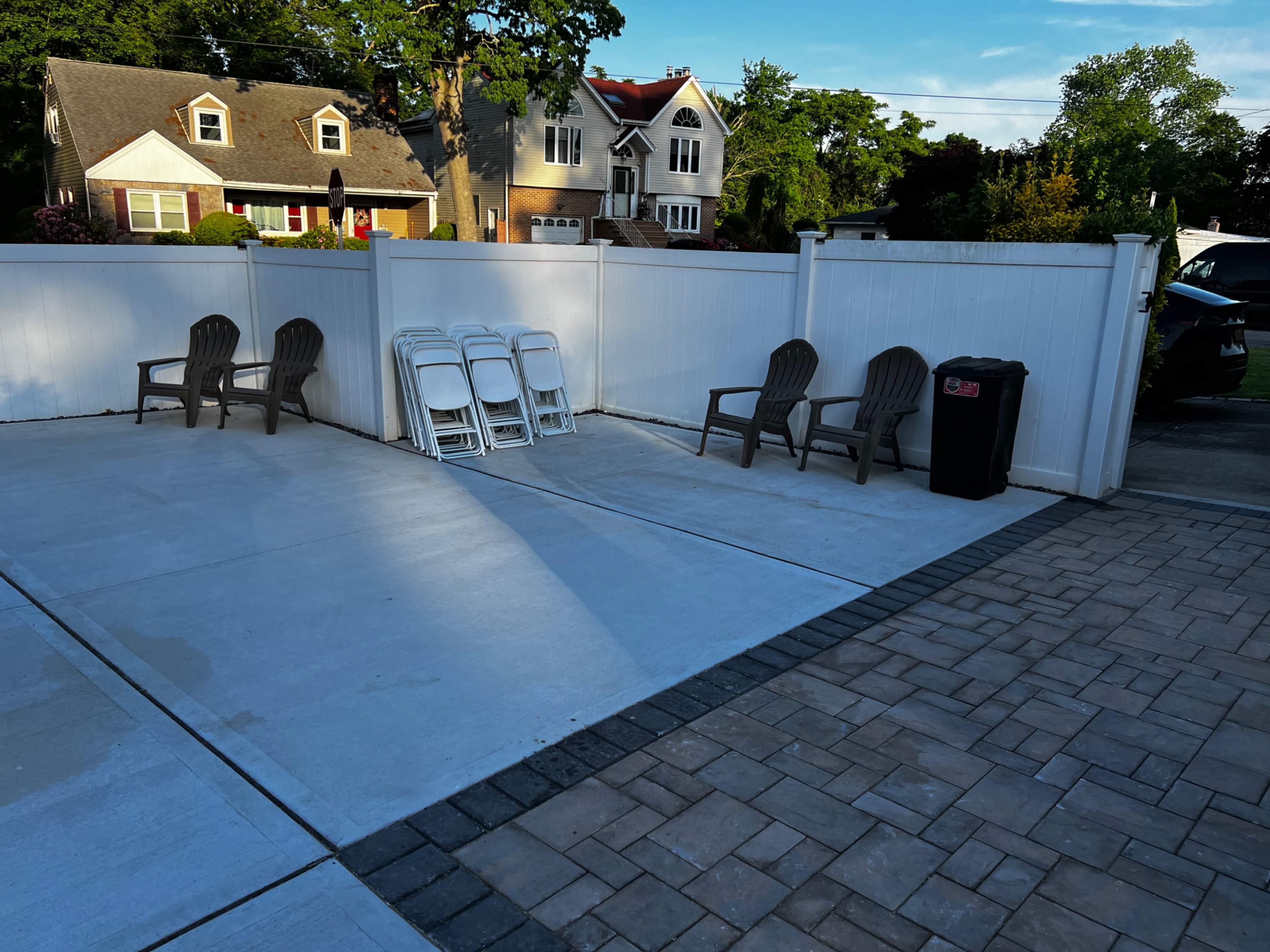 The image shows a paved outdoor area with four plastic chairs, a stack of white folding chairs, and a black garbage bin against a white fence.
