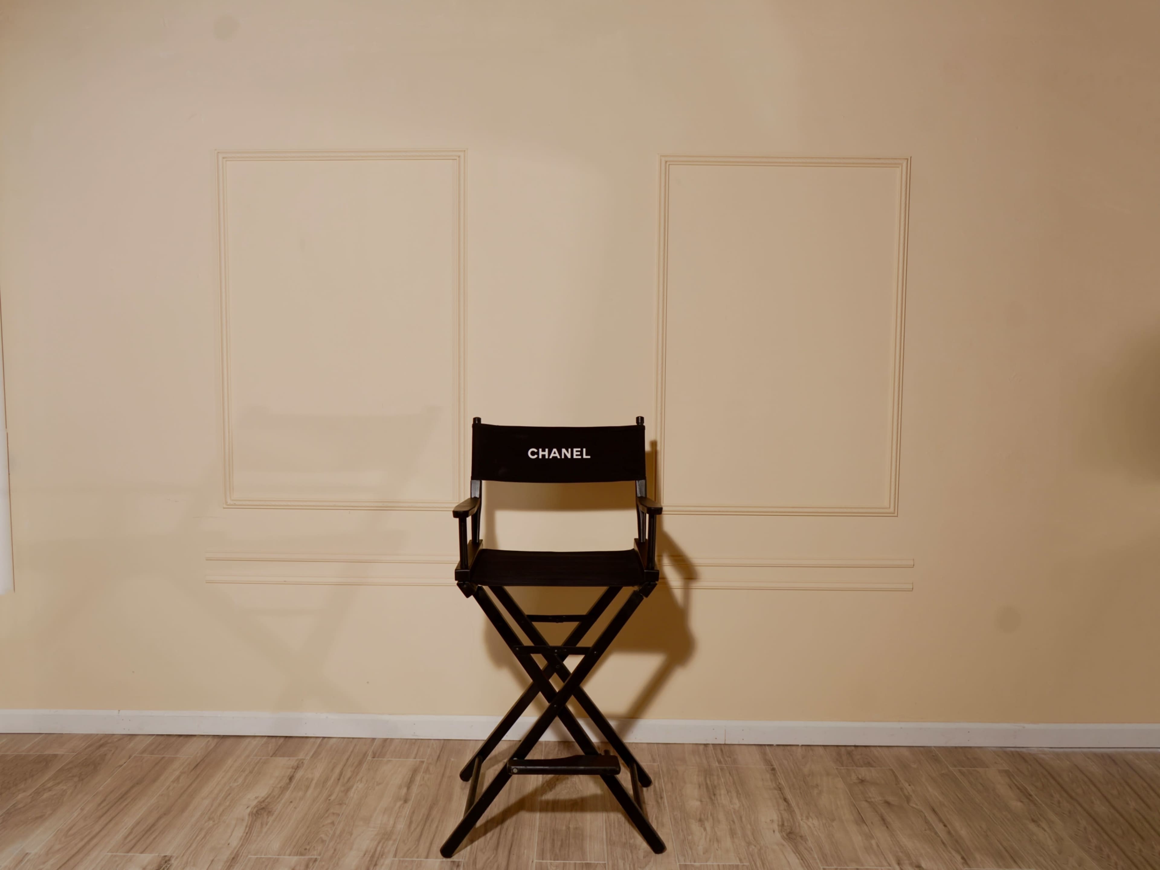 A black director's chair with the name "CHANEL" is positioned against a plain, light-colored wall.