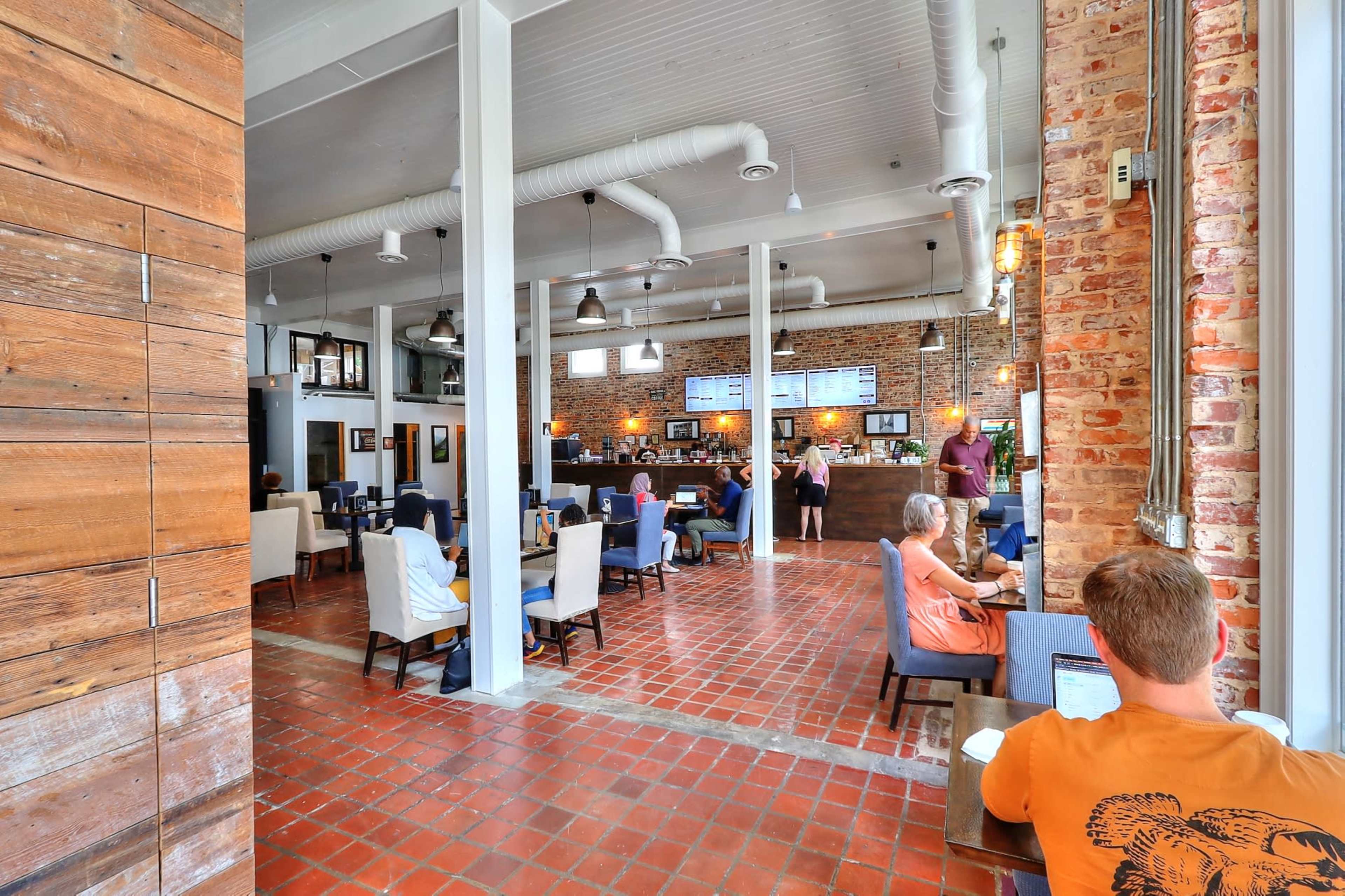A spacious café with a mix of wooden and brick walls, featuring various seating arrangements and a counter in the background where staff interact with customers.