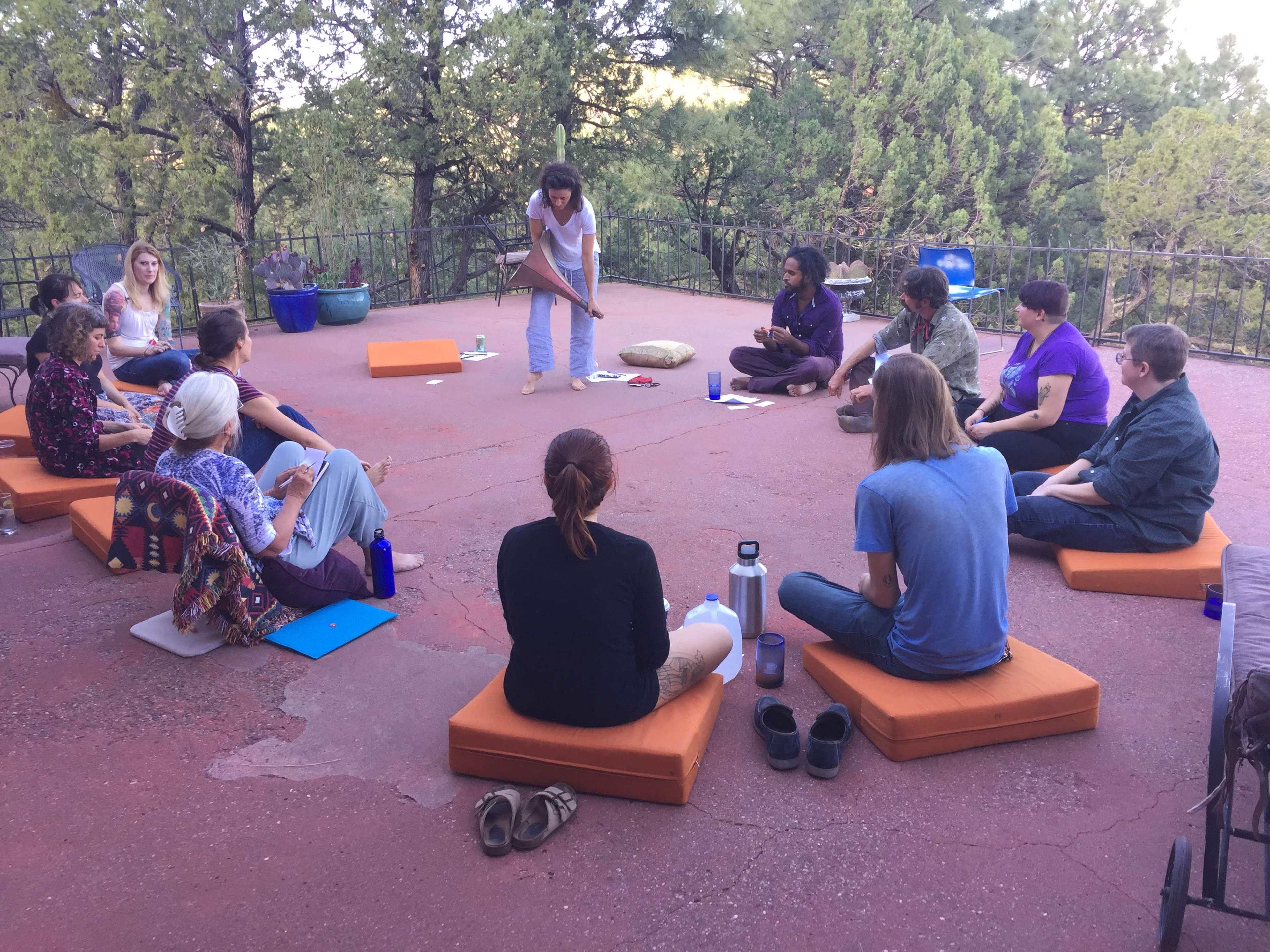 A group of people is seated on cushions in a circle outdoors, engaged in a discussion led by a woman standing in the center.