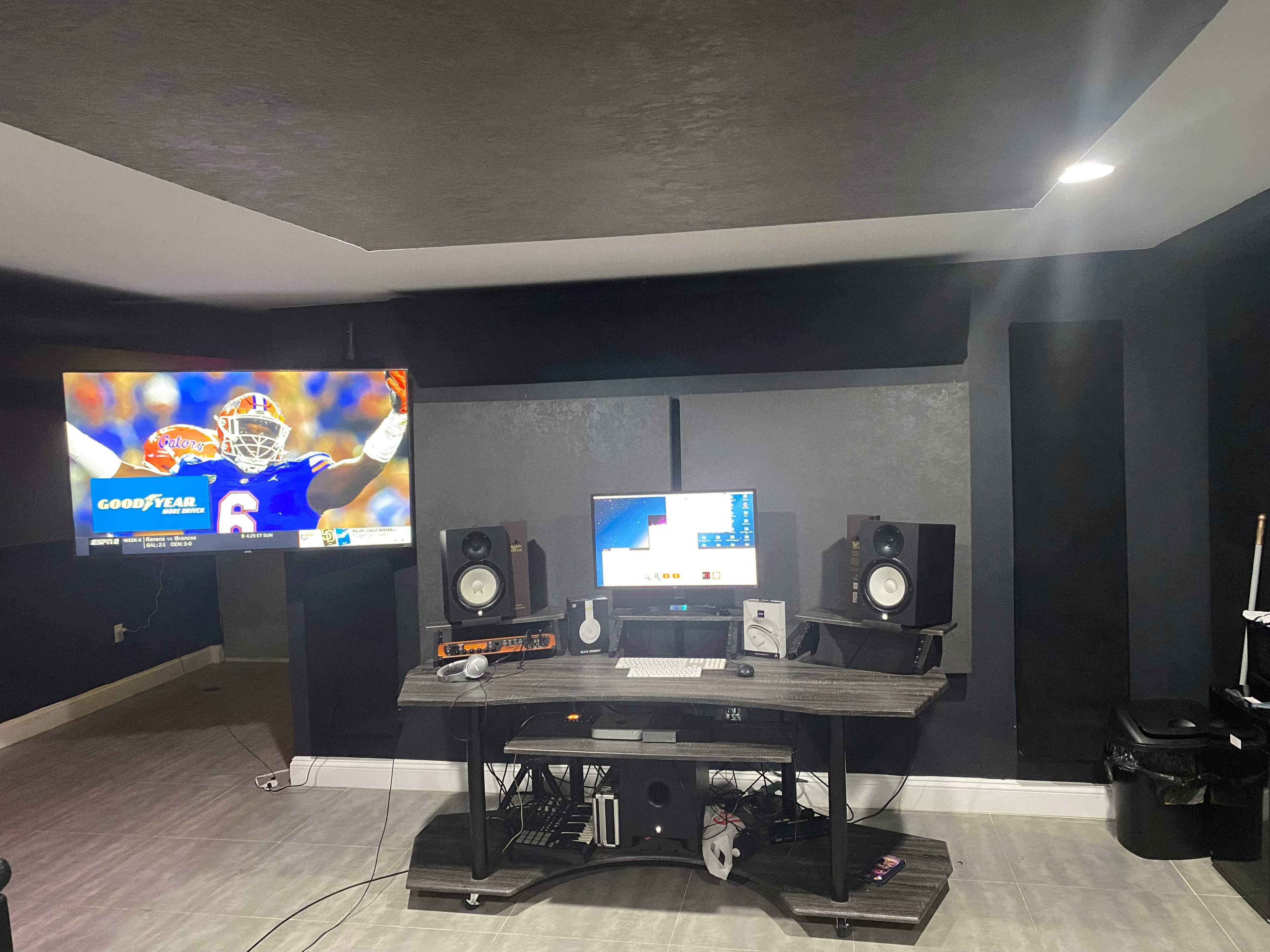A music production or recording studio setup with a large television displaying a sports game, a desktop computer, and studio speakers on a table beneath the screen.