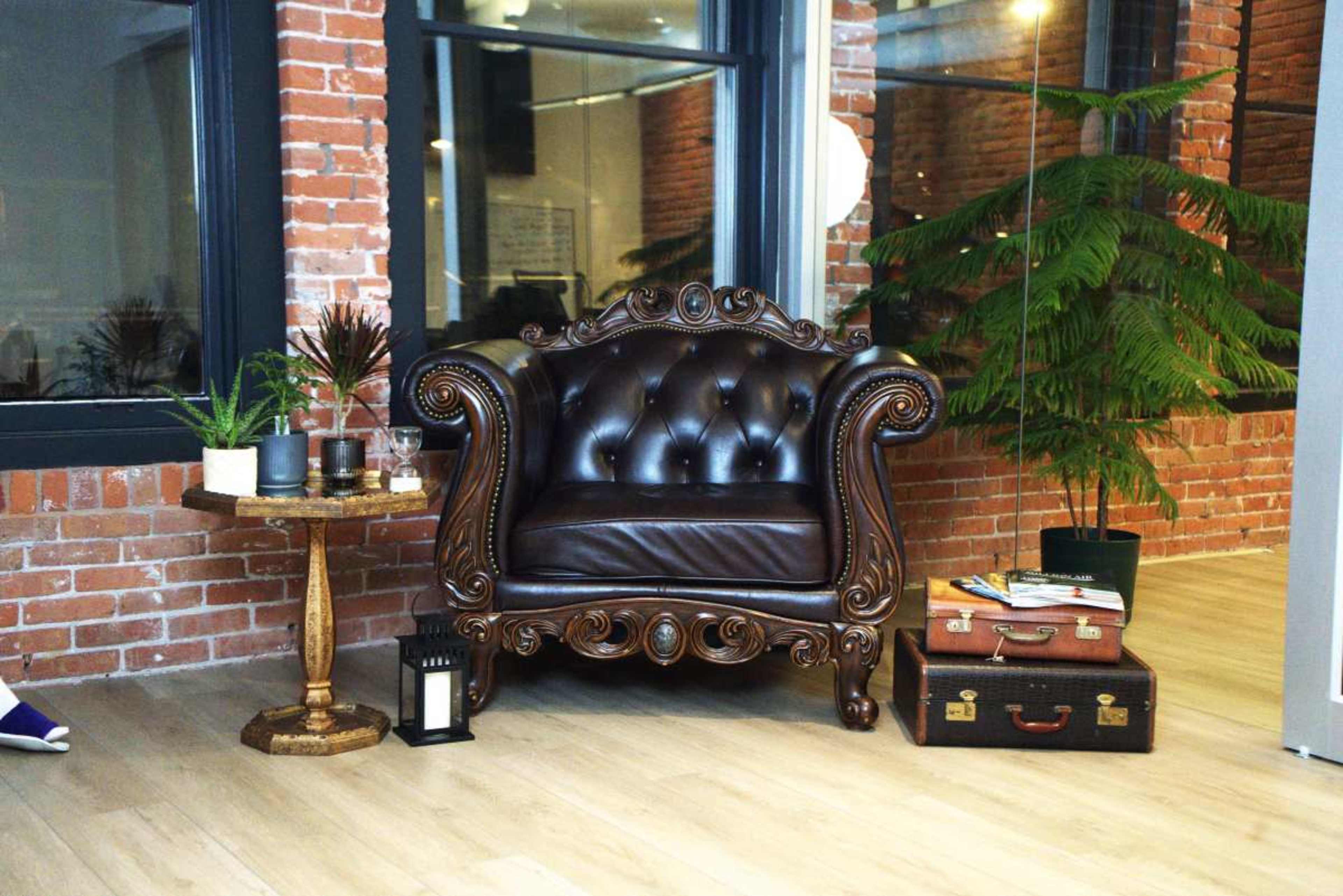 A brown, antique-style leather chair is positioned next to a round wooden side table with plants and a vintage suitcase on the floor in a brick-walled room.