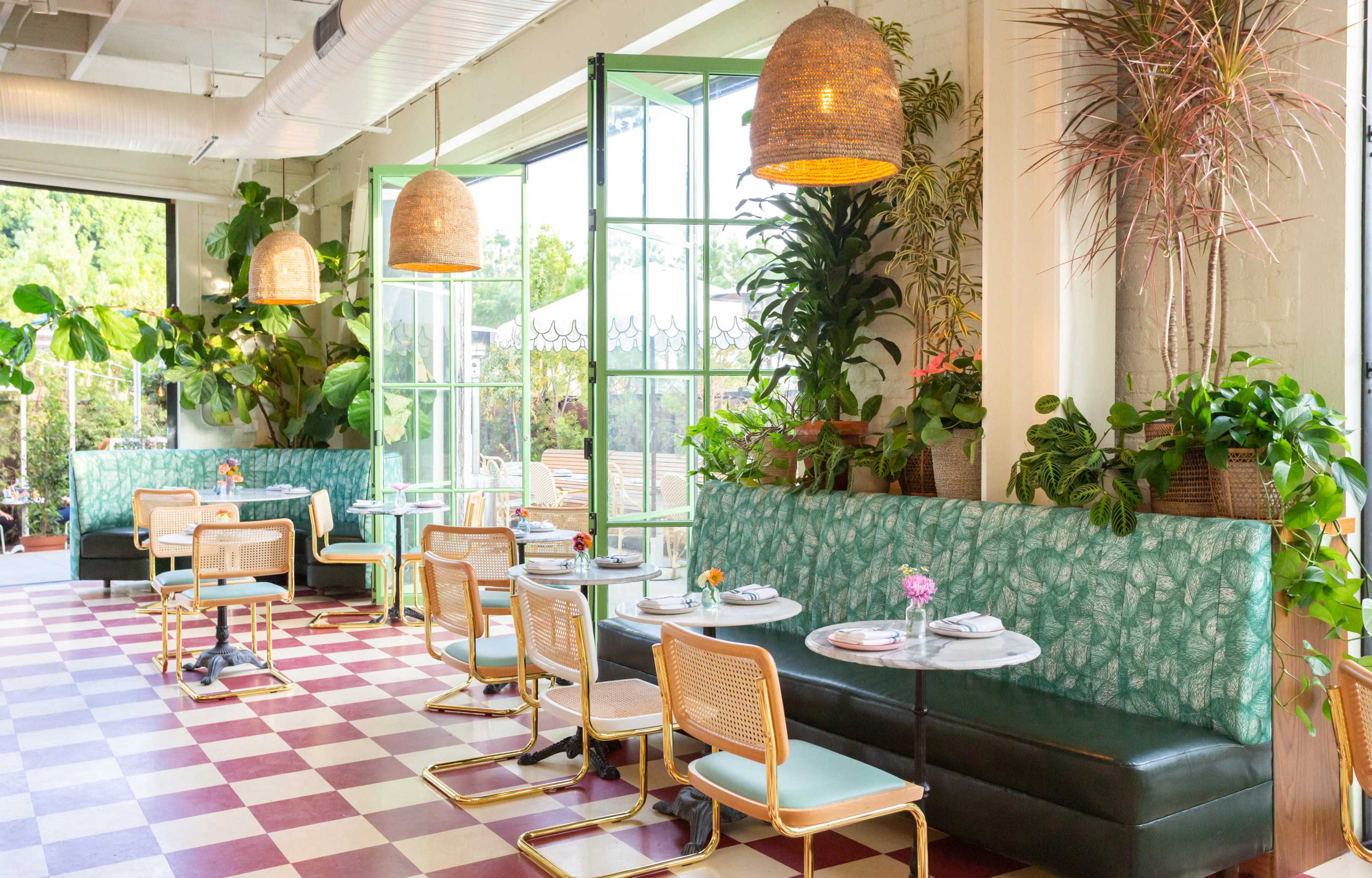 The image shows a brightly lit café with green-potted plants, patterned seating, and round tables arranged on a checkered floor.