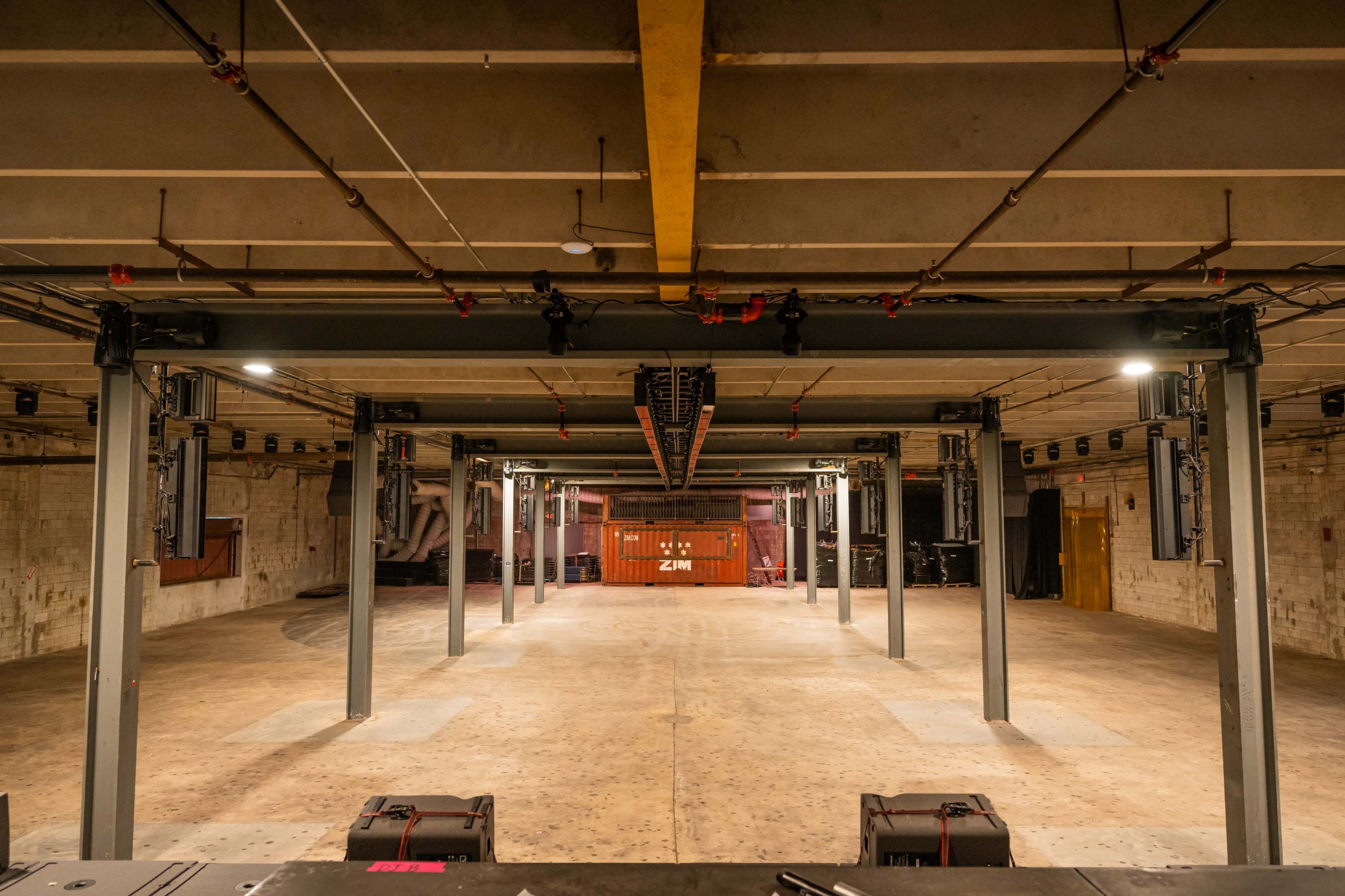 The image shows an empty industrial space with metal columns, overhead lighting, and equipment at the far end, suggesting it is set up for an event or performance.