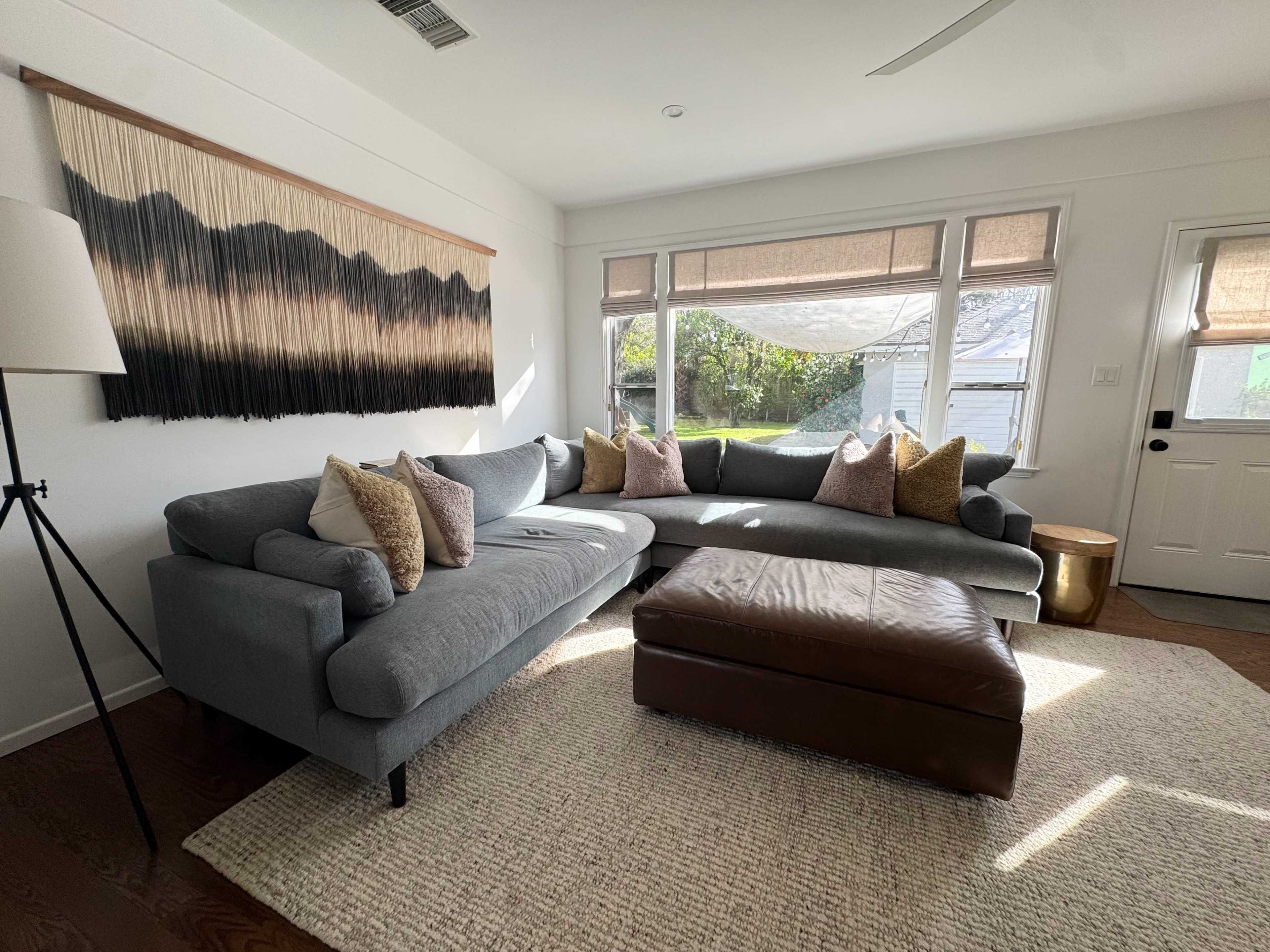 A gray sectional sofa with decorative pillows is positioned in a well-lit living room alongside a brown ottoman and large windows with woven shades.