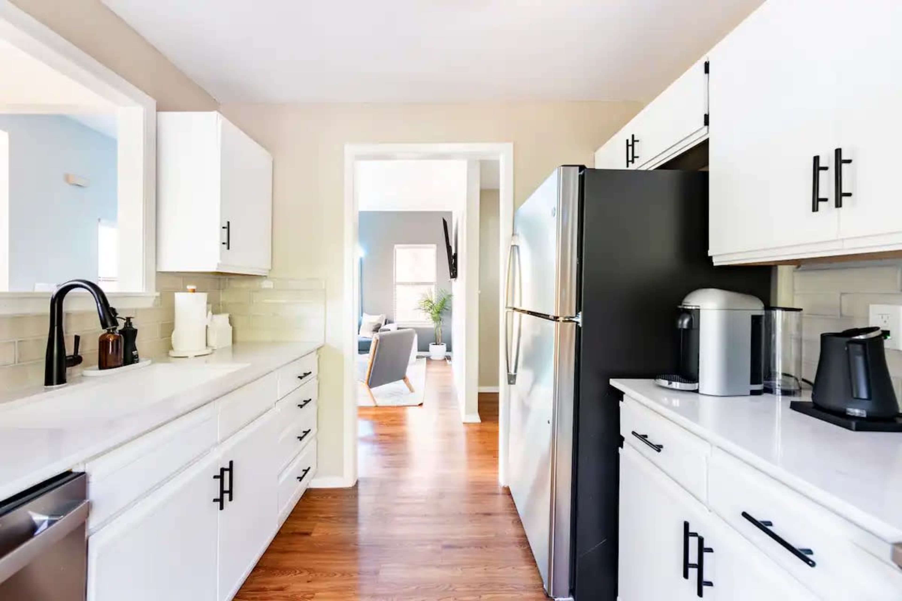 A modern kitchen features white cabinetry, a stainless steel refrigerator, and a view into a light-filled living area.