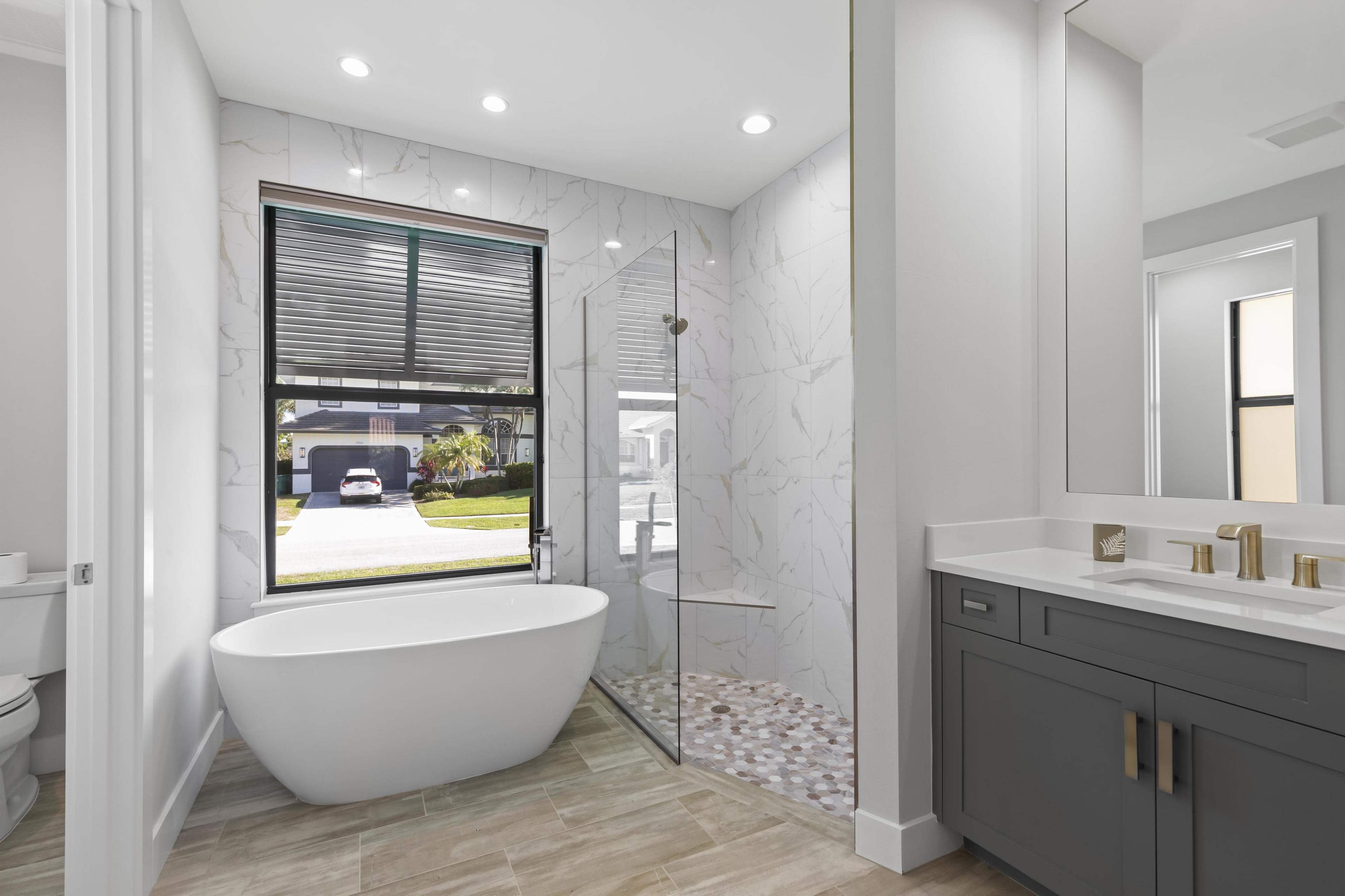 A modern bathroom featuring a freestanding bathtub, a glass shower enclosure, and a dual sink vanity, all set against a marble-patterned wall and a large window.