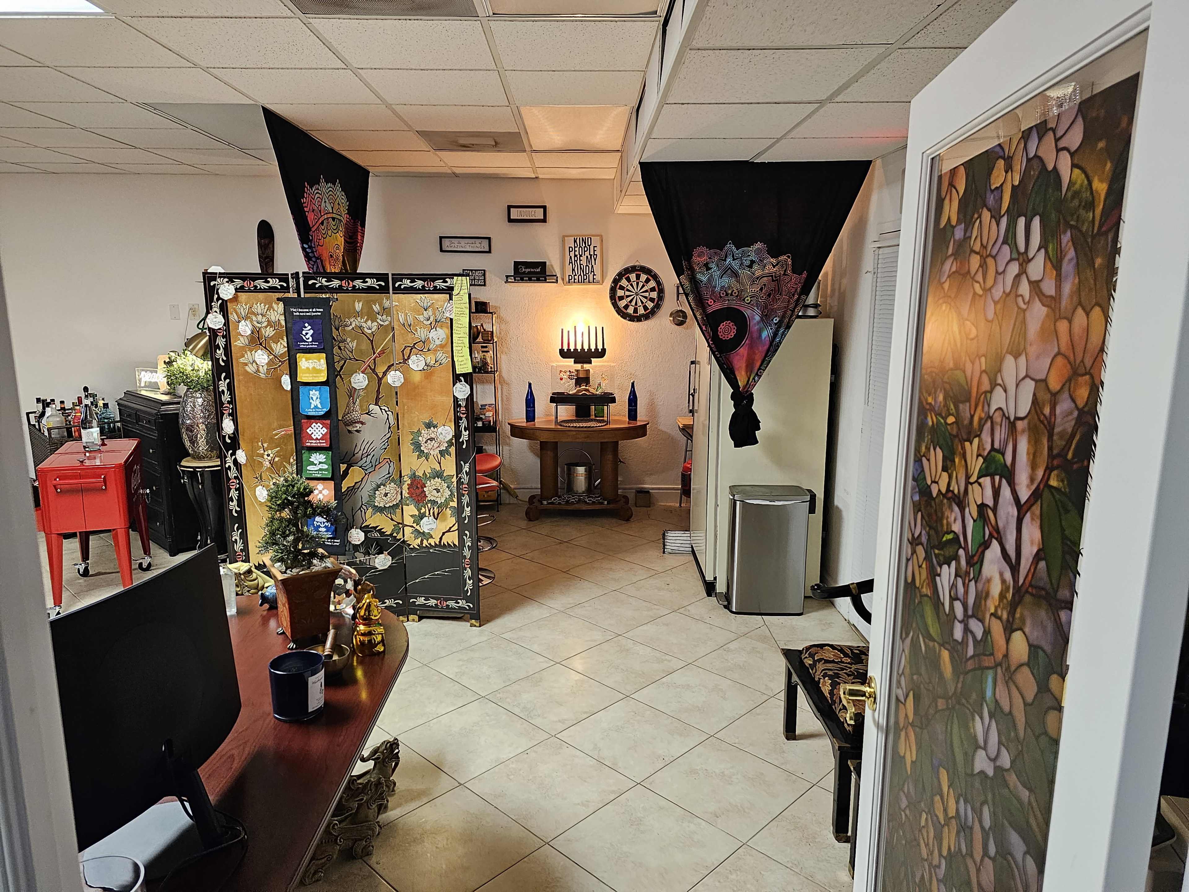 The image shows a room with a mix of decorative elements, including a partition with floral designs, lit candles on a table, and artwork on the walls, alongside a desk and various furnishings.