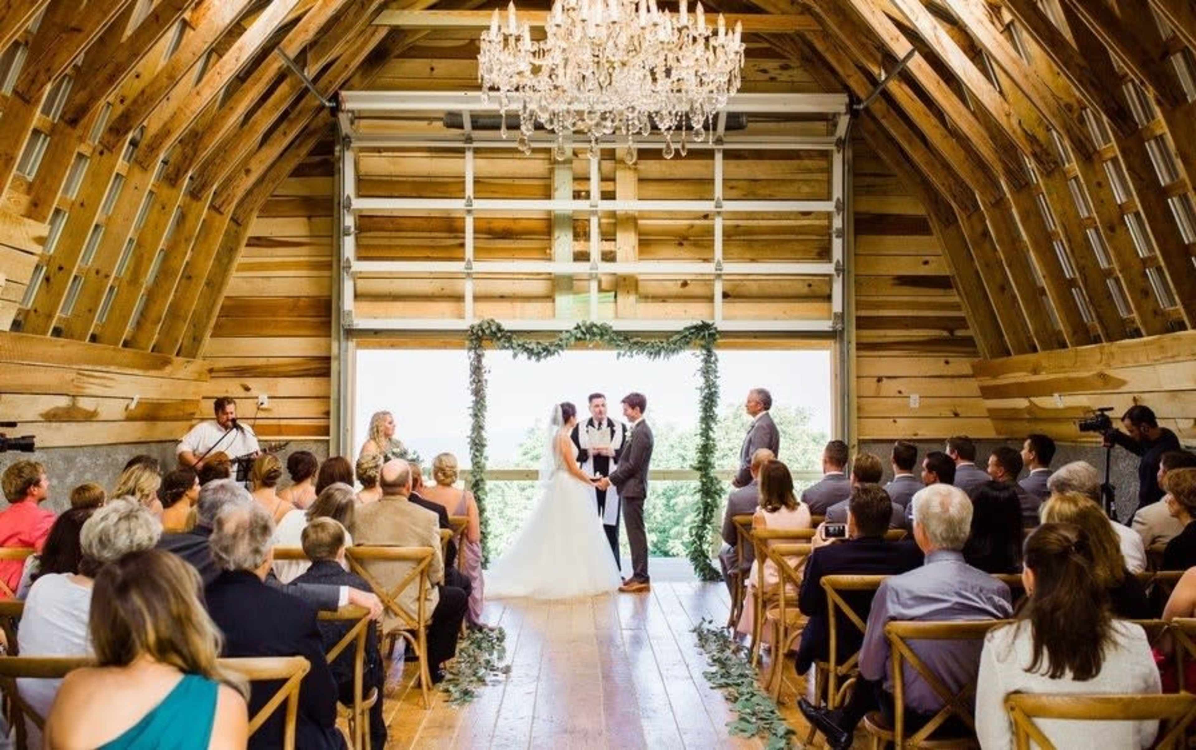 A couple stands at the altar in a rustic barn, surrounded by guests seated on wooden chairs, while a musician plays nearby.