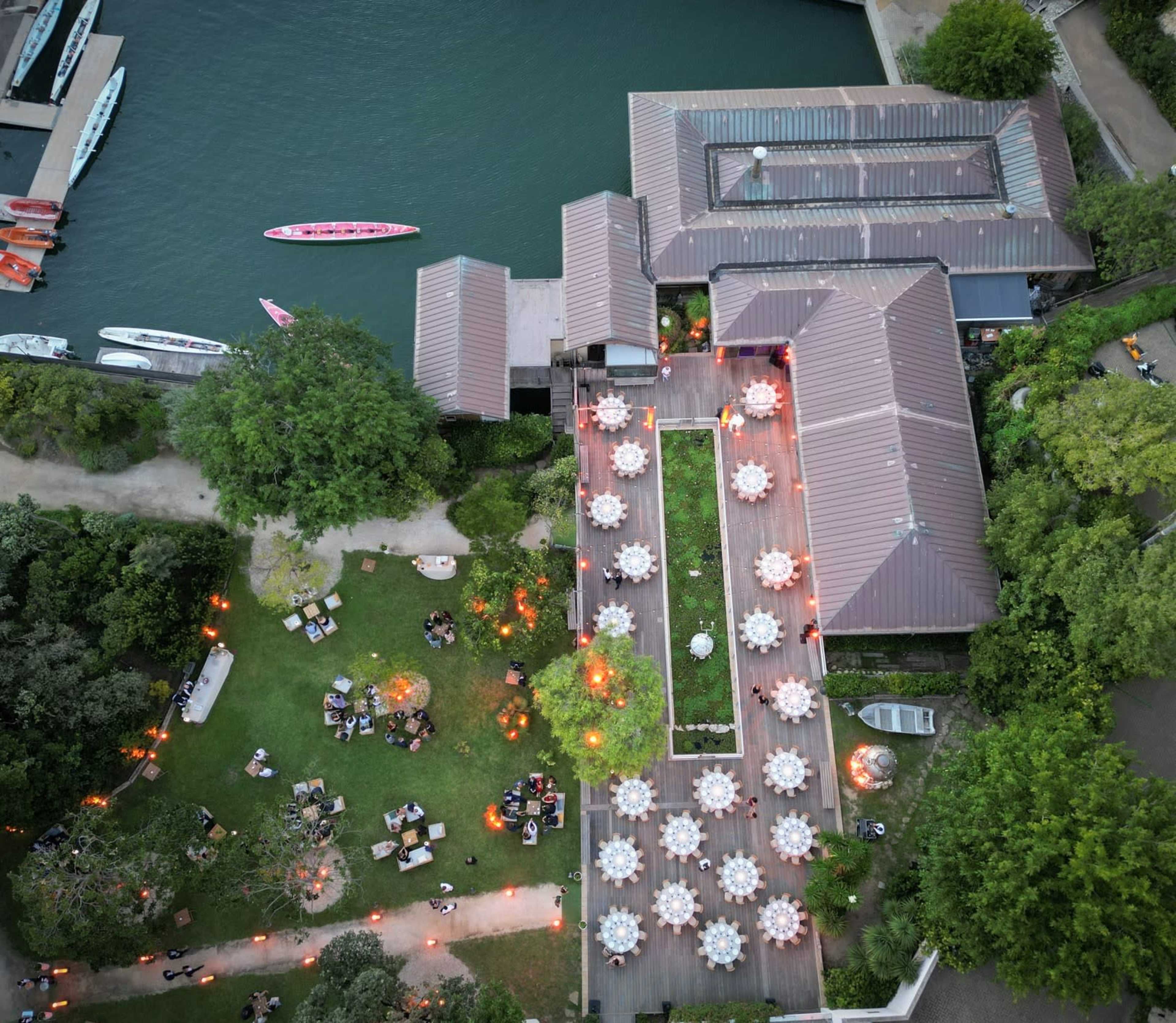 An aerial view shows a riverside venue with multiple tables set up for dining, surrounded by greenery and dotted with soft glowing lights.