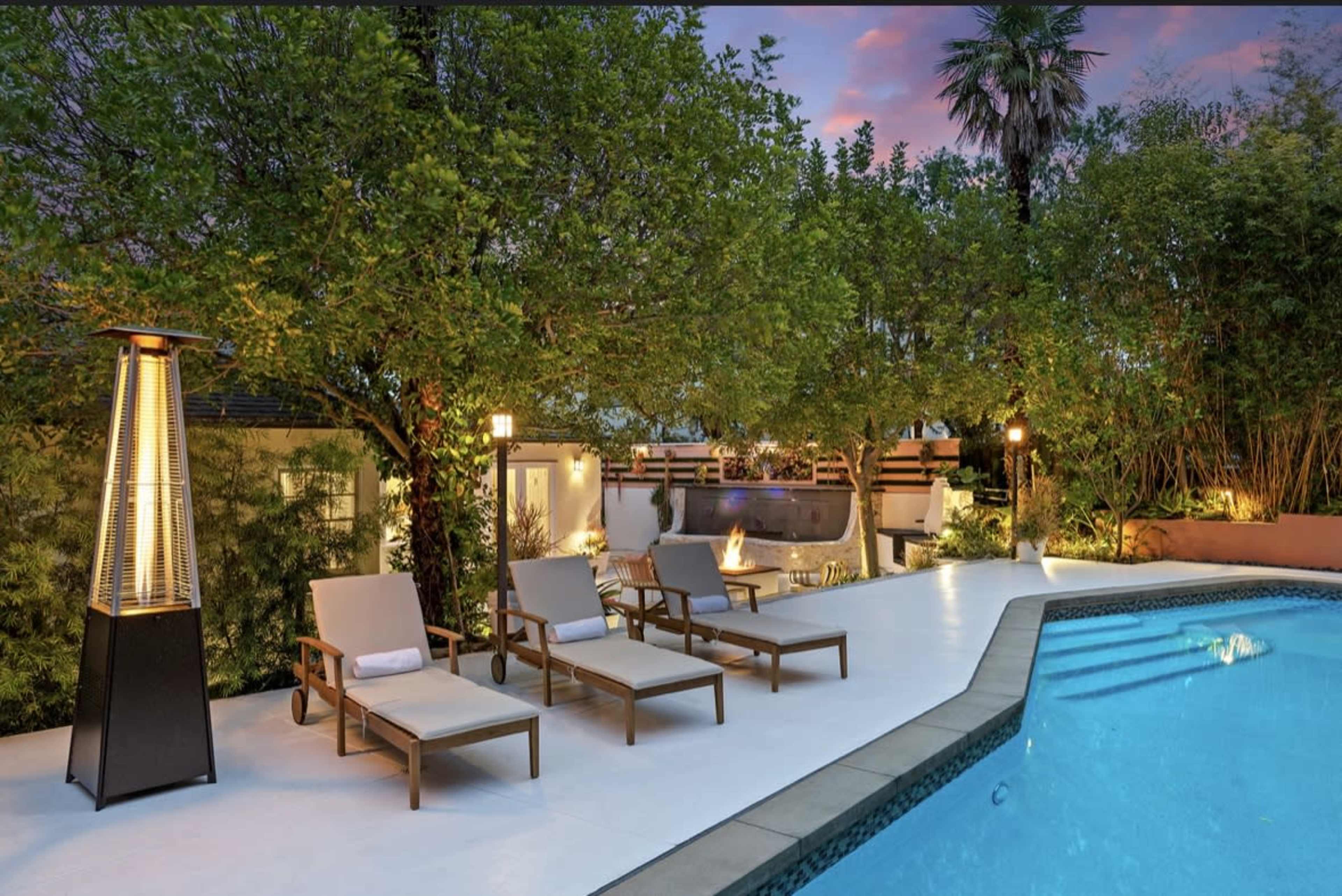 A neatly arranged poolside area features three lounge chairs beside a blue swimming pool, surrounded by lush greenery and illuminated by soft lighting.